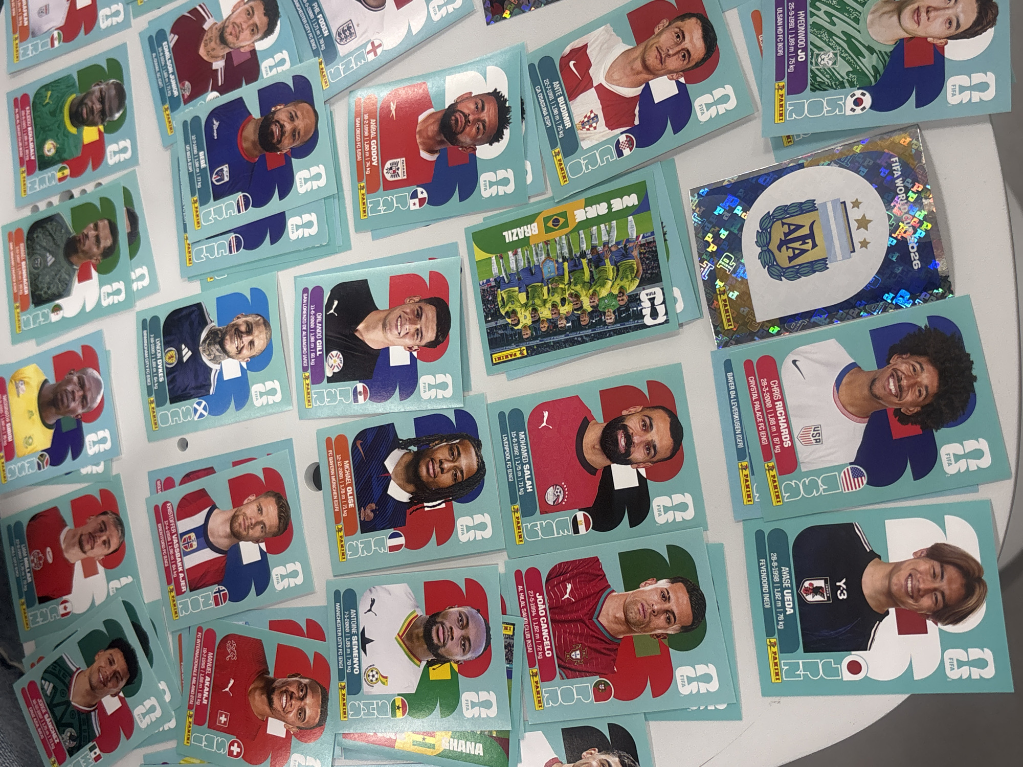 A large collection of Panini football (soccer) trading cards is spread out on a light-colored tabletop in San Andrés Cholula, Mexico. The cards predominantly feature individual male football players, each displaying their portrait in a team or national jersey, along with their name, birthdate, height, weight, and club affiliation.
Visible players and their associated teams or countries include Phil Foden (Manchester City FC, ENG), Mohamed Salah (Liverpool FC, ENG), João Cancelo (Al Hilal Saudi Club, KSA/POR), Manuel Akanji (FC Internazionale Milano, ITA/SUI), Chris Richards (Crystal Palace FC, ENG/USA), Ayase Ueda (Feyenoord, NED), and Anibal Godoy (San Diego FC, USA/PANAMA).
In addition to individual player cards, there is a card featuring the full Brazilian national team squad with the text "WE ARE BRAZIL," and a special holographic card showcasing the Argentina national team crest ("AFA") with "FIFA WORLD 2026" written on it. Each card is bordered in a light blue hue and includes "FIFA" and "PANINI" branding. The cards are arranged casually, suggesting they are part of a collection being viewed or organized. The setting is indoors under artificial light.