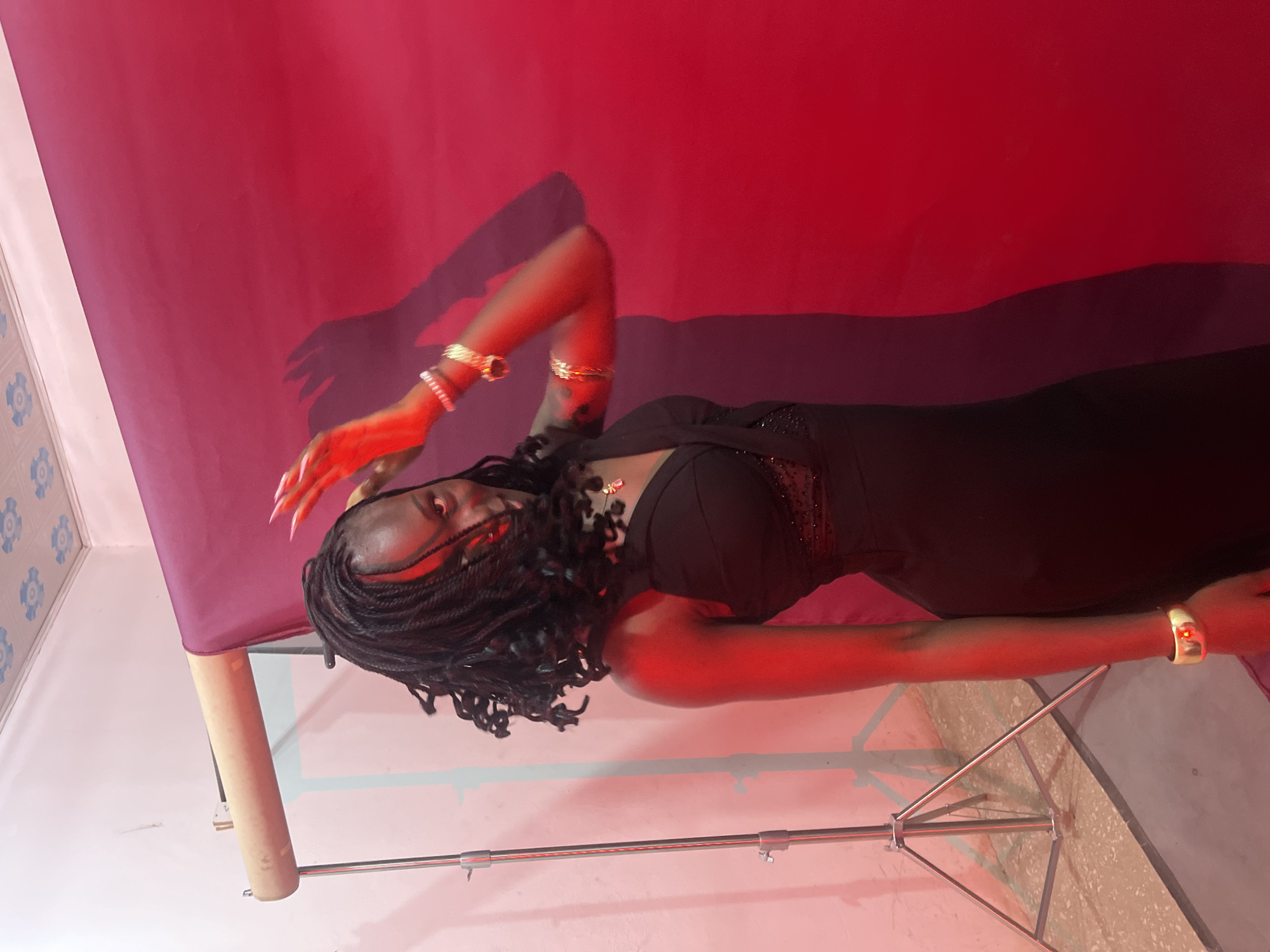 A young Black woman with dark braided hair is posing in front of a red backdrop. She is wearing a black dress and jewelry. The lighting is red, casting shadows on the wall behind her. A portion of a photography stand is visible to the left of the woman. The ceiling appears to be tiled. The floor is concrete with some gravel. The image was taken indoors. The location is Gura Top, Nigeria.