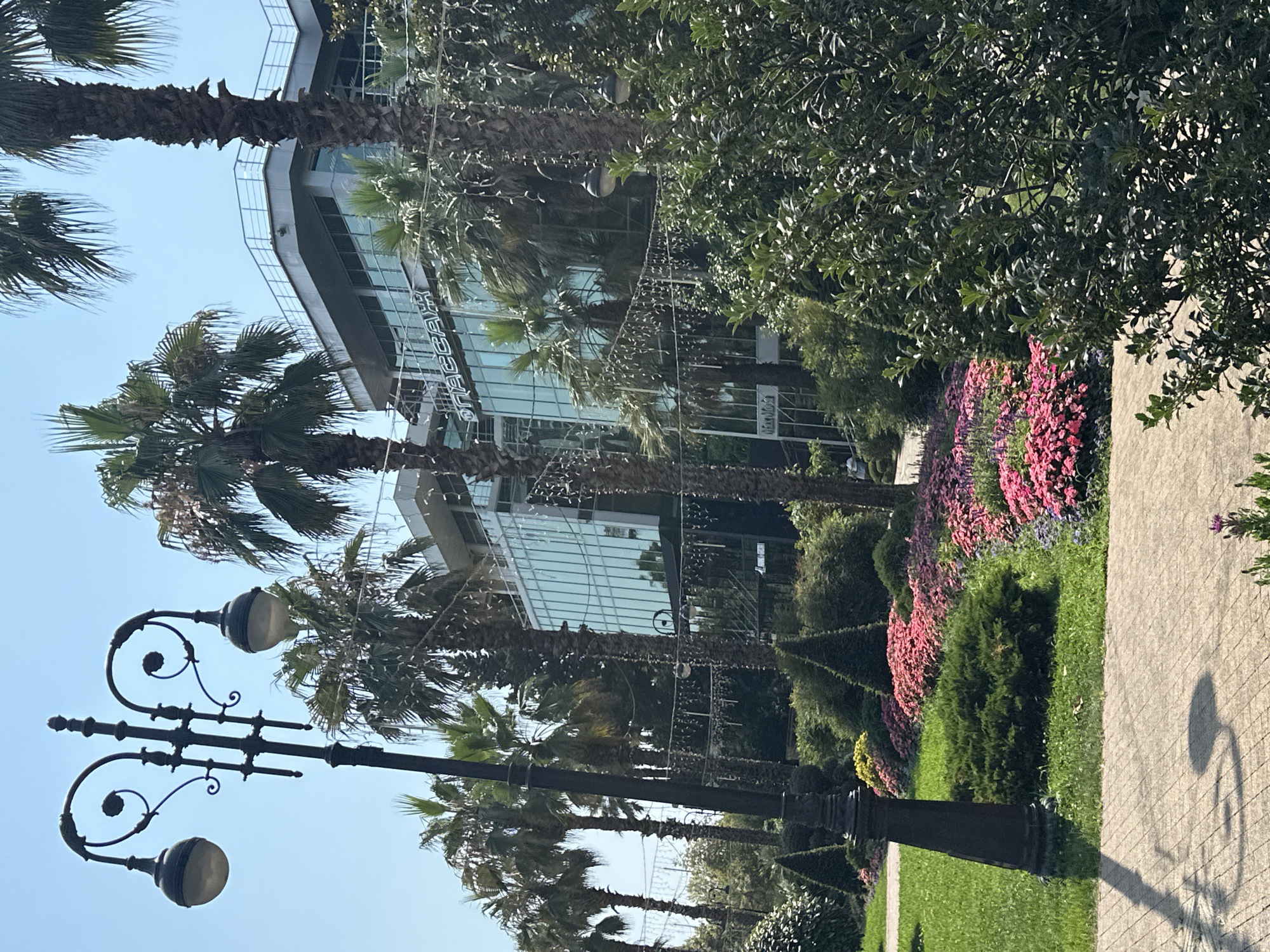 The image shows a sunny day in Sochi, Russia, with a modern glass building and palm trees in the background. In the foreground, there is a lamppost and a path leading through a garden with colorful flowers and manicured bushes. The building, labeled "PASSAGE" in Cyrillic lettering, appears to be a commercial establishment, possibly a shopping center or hotel, given its multi-story structure and extensive glass facade. The lush greenery and palm trees suggest a warm, subtropical climate typical of Sochi. The scene is calm and idyllic, with no visible people actively engaged in any particular activity, though a few figures can be faintly seen in the distance near the building's entrance.