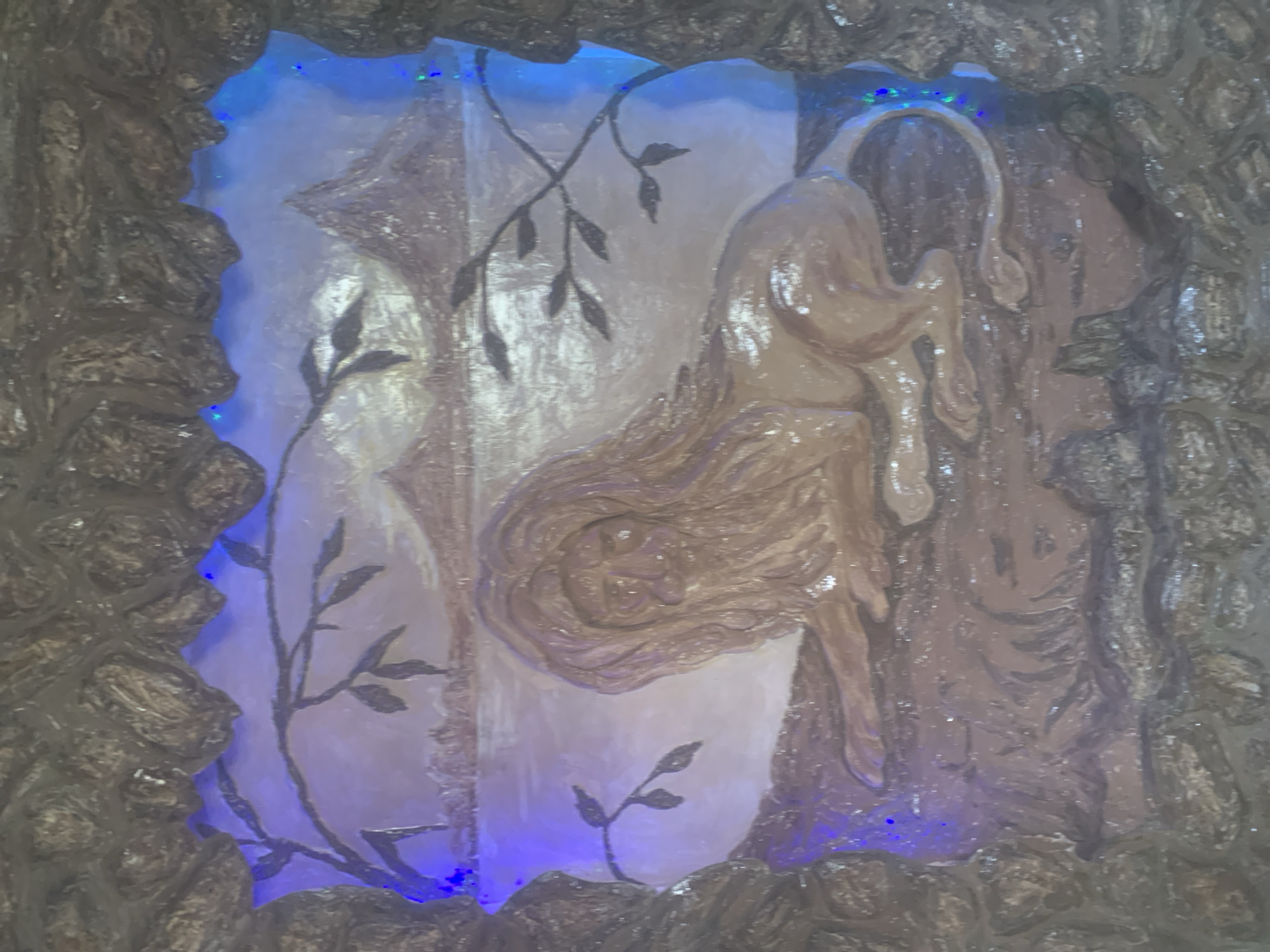 The image shows a bas-relief sculpture framed by a rough, stone-like border. The sculpture depicts a lion reclining in the foreground, with a body of water and mountains in the background. The lion is rendered in a light brown hue, with a flowing mane and tail. The background scene appears to be a landscape with a pale sky and hints of trees or branches. The overall impression is of a stylized, possibly ancient, artwork. The scene is illuminated by blue and green lights, which are visible in the upper part of the frame and along the edges of the sculpture. The stone-like border is textured and appears to be part of the overall installation. It is not possible to determine the exact location within Gyumri, Armenia, based solely on this image.