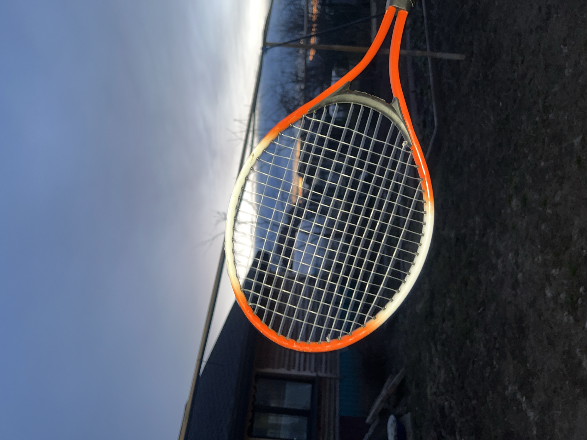 The primary subject of the image is a tennis racket, positioned vertically and prominently in the frame. The racket's frame is a vibrant orange, fading into a lighter, almost white, section towards one side of the head. The strings are metallic silver, forming a dense, regular grid across the racket's face. The grip area, at the top of the visible racket, is dark.
The background is divided between a sky and a landscape featuring structures and ground. The upper portion of the image displays a sky that transitions from a muted blue-grey on the left to a much brighter, almost white hue on the right, suggesting either dawn or dusk with a strong light source just beyond the frame.
Behind the racket, various elements of a rural or suburban environment are visible through its strings. To the left, a long, reflective structure, possibly a greenhouse or a building with a shiny roof, is discernible, reflecting light or distant features. Below the racket's head, a dark building with visible windows, resembling a house or shed, is partially seen. The lower right area of the image reveals dark, textured ground, possibly bare earth or sparse foliage.
No people are present in the image, and no discernible text is visible on the racket or in the background. The image captures an outdoor scene, likely in Votkinsk, Russia, during the soft, atmospheric light of twilight.