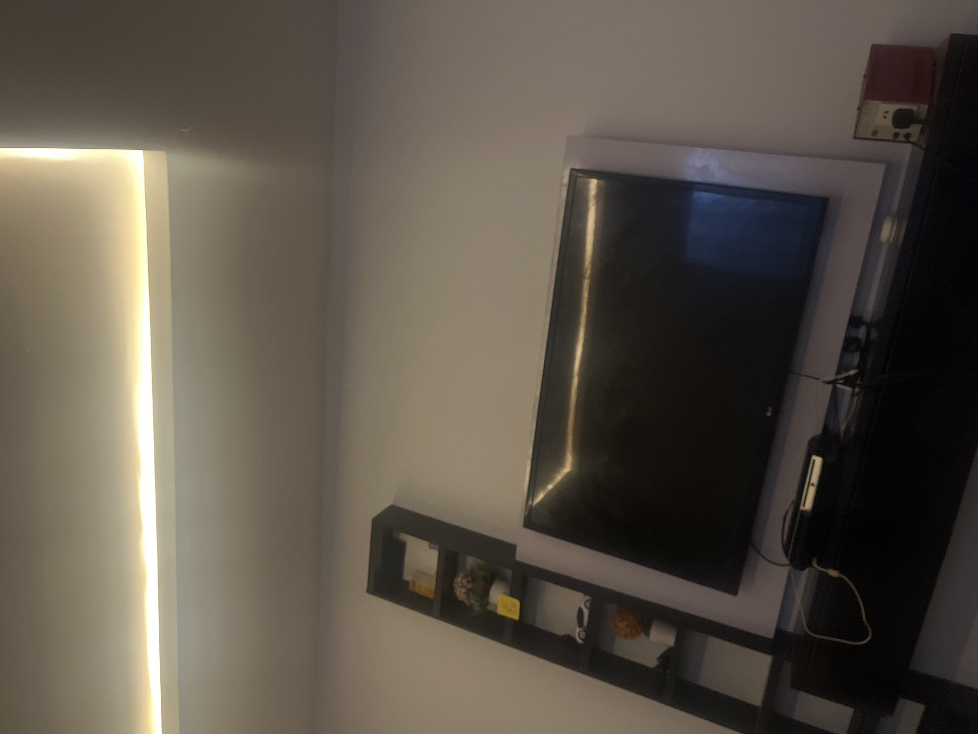 A television is mounted on a wall, with a soundbar beneath it. To the left of the television is a tall, narrow bookshelf with several items on it, including decorative plants and what appears to be a toy car. The room is lit by a recessed ceiling light that emits a warm glow. The walls are painted a light color, and the overall impression is one of a modern, uncluttered living space. The image is taken from a low angle, looking upwards towards the ceiling. No people are visible in the image. This scene is located in Nabatum, Nigeria.