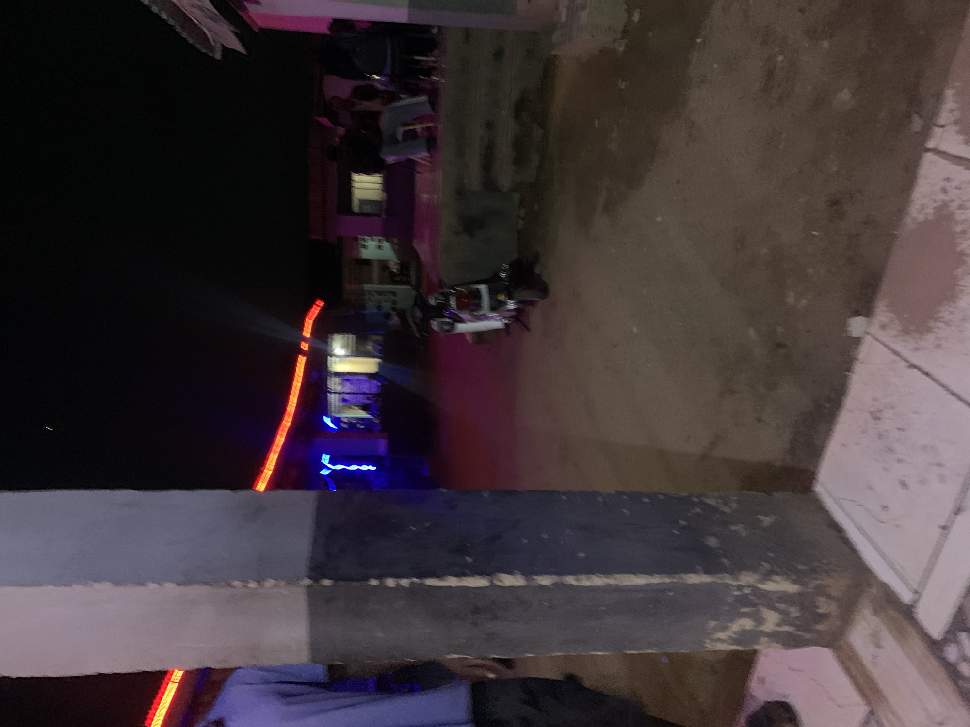 The image shows an outdoor scene in Kabong, Nigeria at night. There are buildings with illuminated blue, red, and white lights. A group of people are visible on a balcony or raised area. A motorcycle is parked on a dirt surface. The foreground features a partially visible structure, likely a wall or a part of a building. Tiles can be seen on the floor. There is a figure near the bottom of the frame, partially obscured.