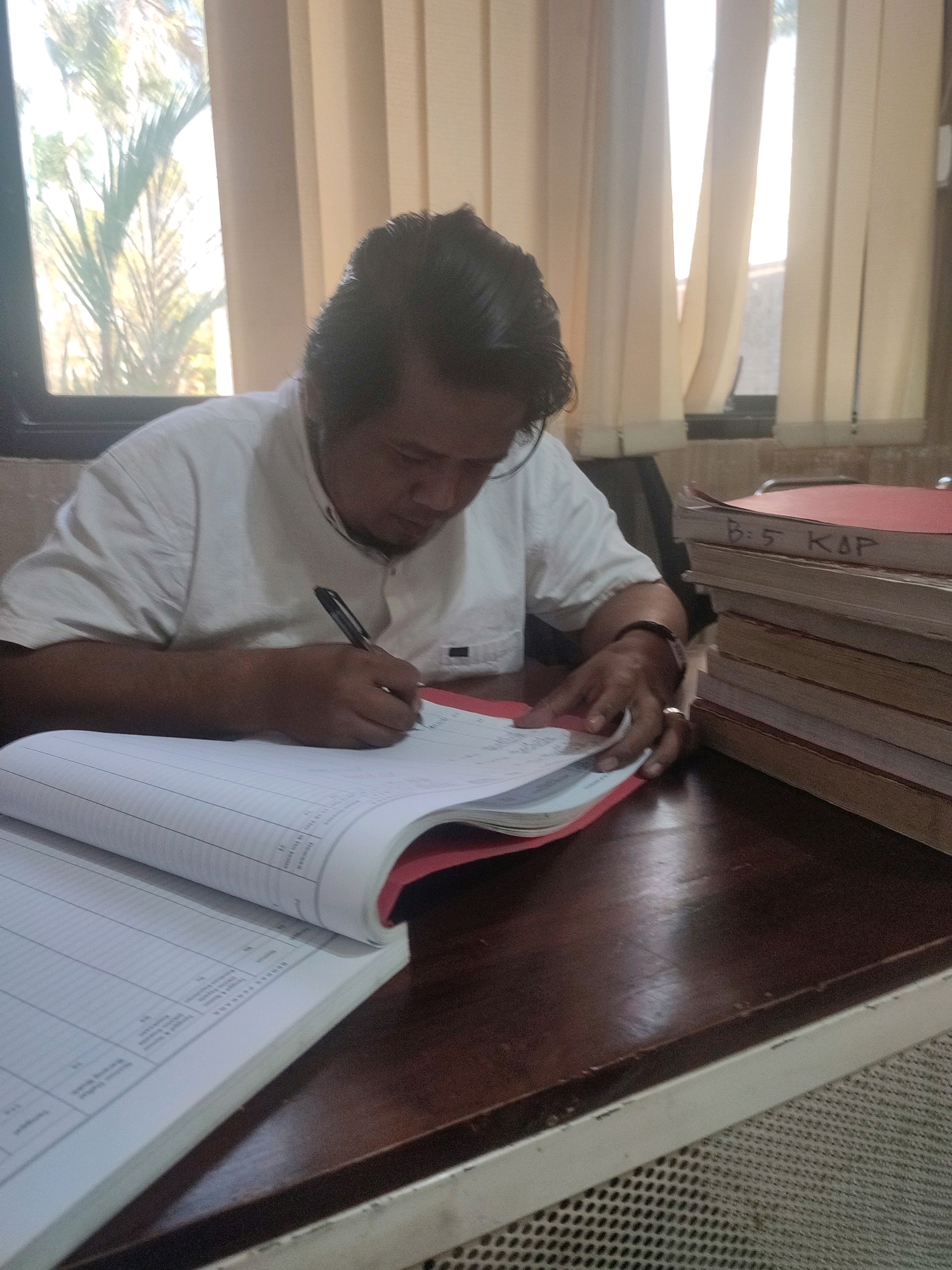A middle-aged East Asian male with dark hair, wearing a white short-sleeved collared shirt, is seated at a dark wooden desk. He is holding a black pen in his right hand and writing in a large, open binder. The binder is filled with lined paper, and appears to be a ledger or record book. To the right of the binder, a stack of red and manila folders is piled high. The folders are labeled with handwritten text. The man is focused on his writing, with his head bowed.
The environment appears to be an indoor office setting. Behind the man, a window is visible with vertical blinds drawn, allowing sunlight to filter through. Outside the window, greenery, possibly palm fronds, can be seen. The desk has a worn, polished surface. The overall lighting suggests it is daytime. The location is identified as Ringinanom, Indonesia.
