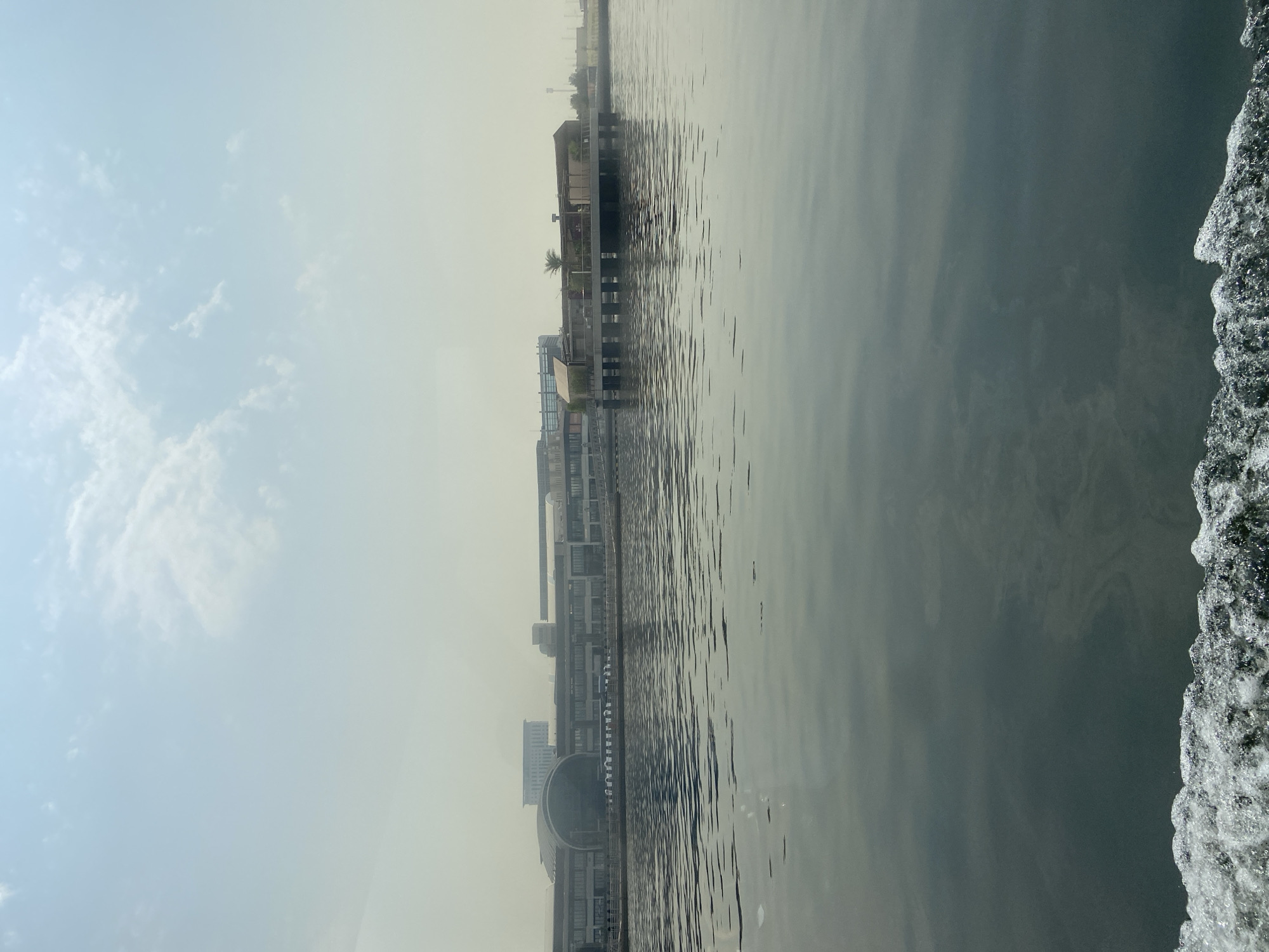This image shows a view from a body of water, likely the Dubai Creek, looking towards a large, modern building complex and a smaller, elevated structure with a palm tree. The sky is hazy and light blue with scattered clouds. The water in the foreground is dark and choppy, with white foam from a boat's wake. The building complex in the background appears to be a commercial or entertainment venue, with large windows and a distinctive arched entrance. The elevated structure to the right seems to be a restaurant or cafe, with outdoor seating visible. The overall scene suggests a daytime view of a waterfront area in Dubai, possibly a tourist or recreational district.