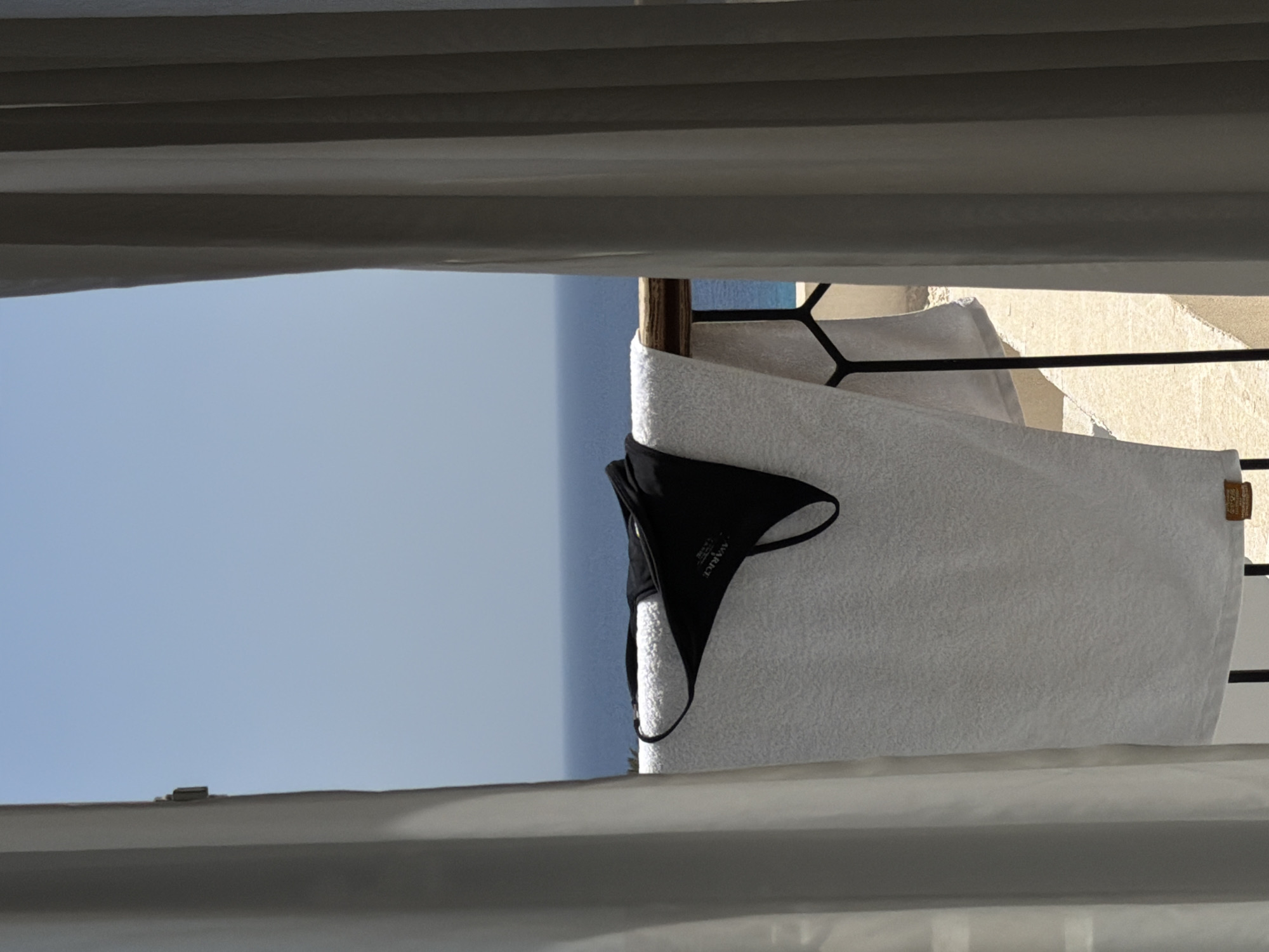 This image captures a bright, sunny scene, likely from a balcony or window overlooking a vast blue expanse. The perspective is framed by grey, horizontal fabric elements, possibly drapes or blinds, at the top and bottom, creating a layered effect.
In the mid-ground, a black bikini top and a white towel are hanging horizontally over a dark metal railing, presumably drying. The bikini top is positioned to the left of the towel and has the brand name "AVARICE" visible in white text on its fabric. The towel is thick and appears soft, extending across much of the visible railing.
Behind the railing, a bright blue sky dominates the view, suggesting clear weather. A distant hint of the sea is also likely part of this blue horizon, characteristic of a coastal location like Sharm El Sheikh, Egypt. To the right of the towel, glimpses of light-colored architecture, possibly a building or wall with textured surfaces, are visible through the gaps in the railing and between the hanging items. The sunlight strongly suggests it is daytime. The overall impression is one of relaxation and vacation in a warm, sunny destination.
