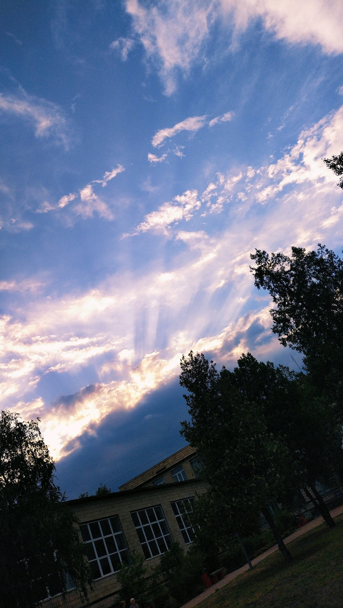 The image captures a dramatic sky over a school building in Horishni Plavni, Ukraine. The sky is filled with a mix of bright, sunlit clouds and darker, shadowed clouds, creating a striking contrast. Rays of light, known as crepuscular rays, pierce through the clouds, illuminating the scene. Silhouetted trees frame the top right and left portions of the image, adding depth and texture. In the lower half of the image, a light-colored brick school building with large, multi-paned windows is visible. The building is surrounded by greenery, including trees and bushes. The overall impression is one of natural beauty and urban architecture coexisting.