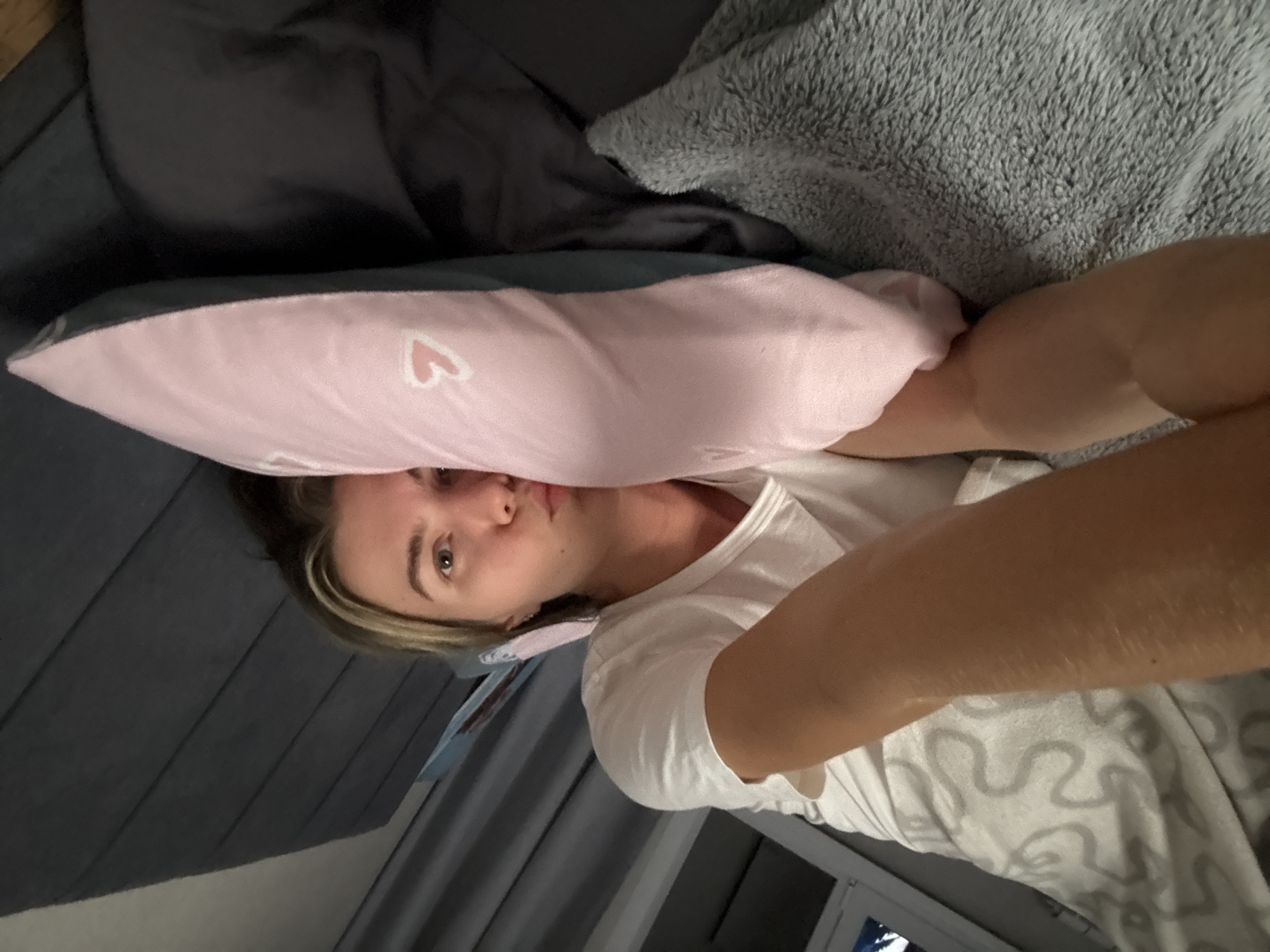 A woman lies on her side on a bed, facing the camera. She is wearing a white t-shirt and is propped up by a pink pillow with white hearts on it, which rests on a dark gray upholstered headboard. To her left, a grey, fuzzy blanket is visible. Her arm is bent, and she is looking directly at the camera with her eyes open. The background shows a window with grey curtains.