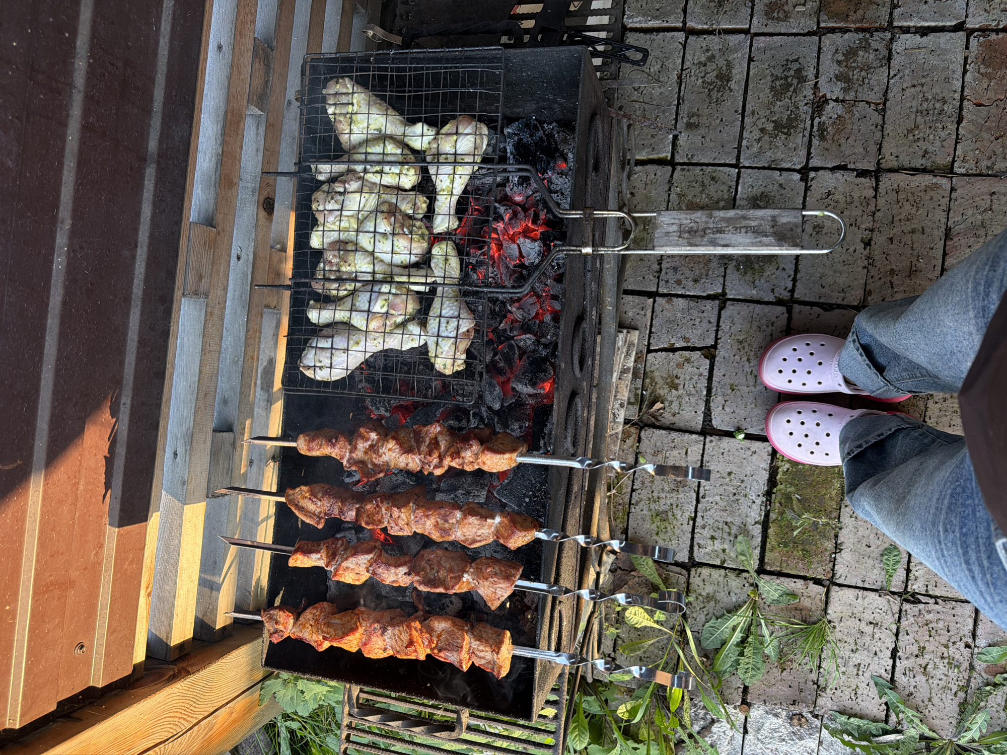 The image captures an outdoor grilling scene in Kamenka, Russia, on a paved brick surface. In the foreground, a pair of light pink Crocs are visible, accompanied by denim jeans. The focus of the image is a charcoal grill, with glowing red embers at the bottom. On the left side of the grill, four skewers of meat, likely shashlik, are cooking. To the right, a wire grill basket holds several chicken drumsticks, also cooking. The background shows a wooden structure and some greenery. The lighting suggests it is daytime, possibly late afternoon due to the warm tones.