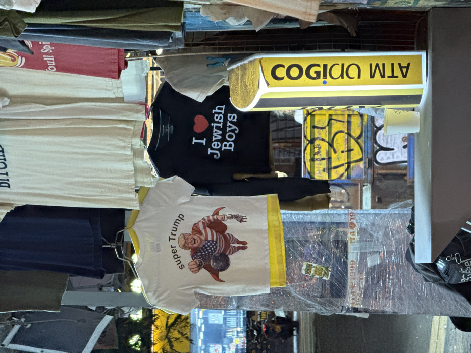 The image displays a nighttime street vendor stall in Tel-Aviv, Israel, exhibiting various t-shirts on hangers. In the foreground, a white t-shirt featuring a cartoon caricature of Donald Trump as "Super Trump" is prominently displayed. He is depicted in a red and white striped cape resembling the American flag, holding a wrecking ball with a blue Star of David on it in his right hand, and a small, caricatured figure by a rope in his left hand. Below this, a black long-sleeved t-shirt reads "I ❤️ Jewish Boys" with a red heart symbol. Other shirts, some partially visible, hang above and around these, including one with the word "BITCHES" visible at the top.
To the right, a vertical, illuminated yellow sign with black text reads "COGIGO" (קוגיגו in Hebrew) and "ATM" (English) arranged vertically. A white paper cup rests on a dark counter surface in front of this sign. To the left, a vertical structure is wrapped in clear plastic sheeting, revealing faded white labels ("ITA 1999") and a green sticker underneath. The background shows blurred elements of a city street, including distant lights from other establishments, yellow wall graffiti, and a parked vehicle on the far left. No people are visible within the frame.