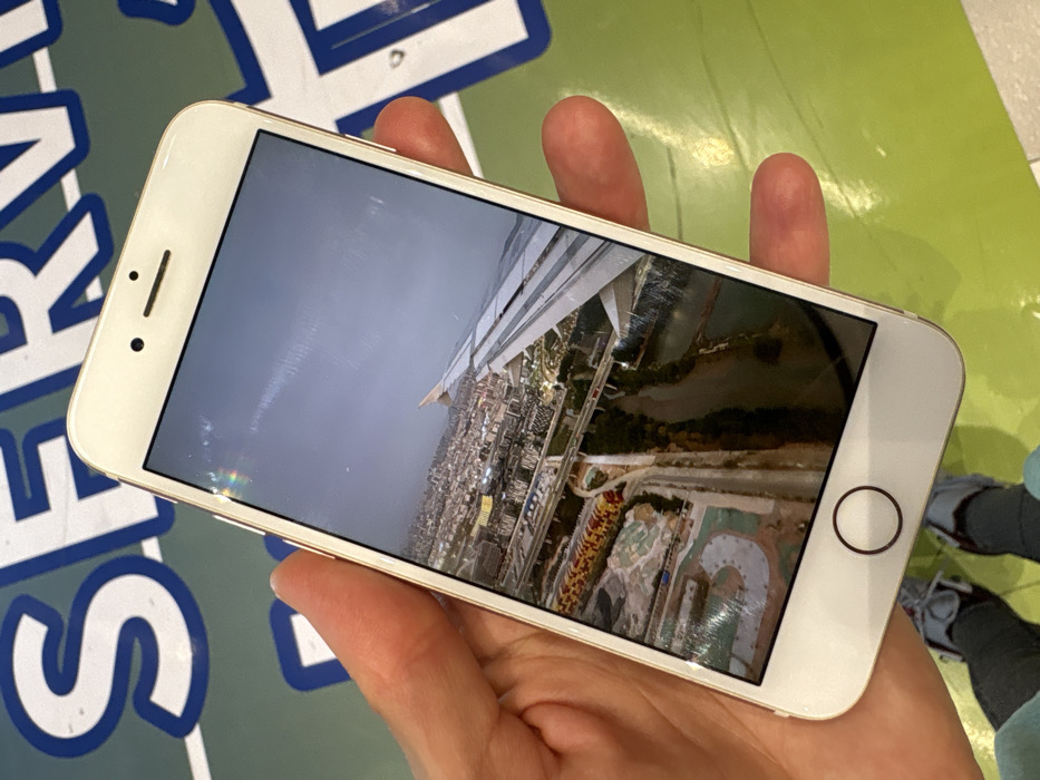 A human hand, appearing light-skinned, holds a white smartphone vertically over a glossy, lime-green surface. The phone's screen displays an aerial photograph, viewed from an aircraft. The image on the screen depicts a partial white aircraft wing with visible flaps extending over a sprawling urban landscape. This landscape features numerous buildings, an extensive network of roads and bridges, a winding river or canal, and a distinctive multi-colored (yellow and red) patterned area. The sky above the urban area is overcast and dim. Below the hand and phone, the lower legs of an individual wearing light blue trousers and grey-and-white sneakers are visible. The green background surface bears large, partial white and dark blue lettering, spelling out "SERV," likely part of a word like "SERVICE." The depicted urban landscape is located in Tianya District, China.