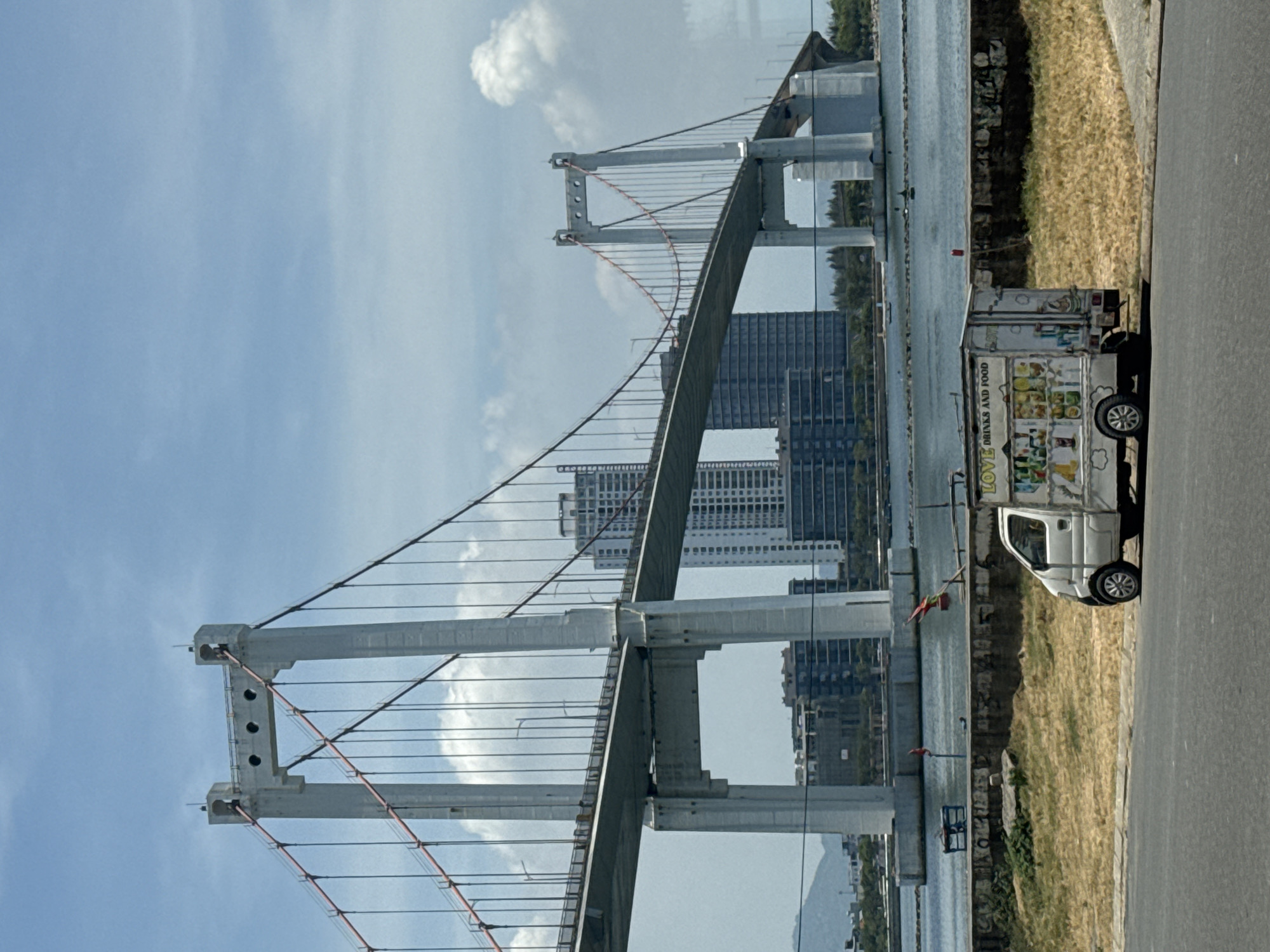 A street-level, eye-level shot shows a bridge in Đà Nẵng, Vietnam. The bridge is a cable-stayed bridge with white support towers. The main span appears to be a concrete arch. The background includes the sky, several high-rise buildings, and water. On the right side of the frame, a white food truck labeled "Love Drinks and Food" is parked on the side of the road. The road itself is asphalt. Vegetation is seen behind the food truck. The overall tone is bright and sunny.