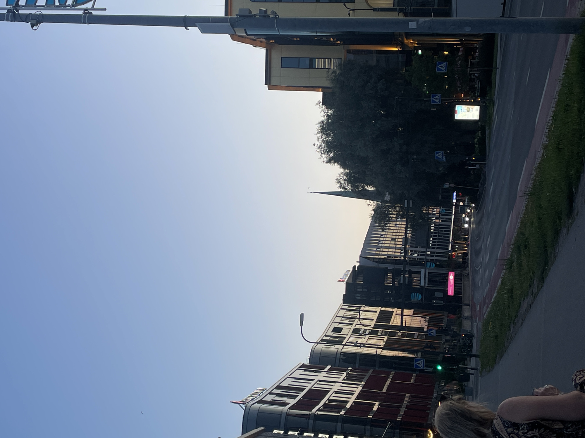 The image shows a street scene in Tallinn, Estonia, during dusk. On the left, a woman with blonde hair is seen from behind, wearing a patterned dress, looking towards the street. She is standing on a sidewalk next to a grassy area. Across the street, there are modern buildings with distinctive architecture. One building on the left has red and dark-colored panels. Further down the street, more buildings with reflective windows are visible. A traffic light is showing green. A church spire can be seen rising above the trees in the background, suggesting the area has a mix of modern and older structures. The sky is clear and a pale blue, transitioning into a lighter hue near the horizon, indicating the time of day. There are streetlights and traffic signs present. Cars are visible on the road, and a digital billboard is lit up.