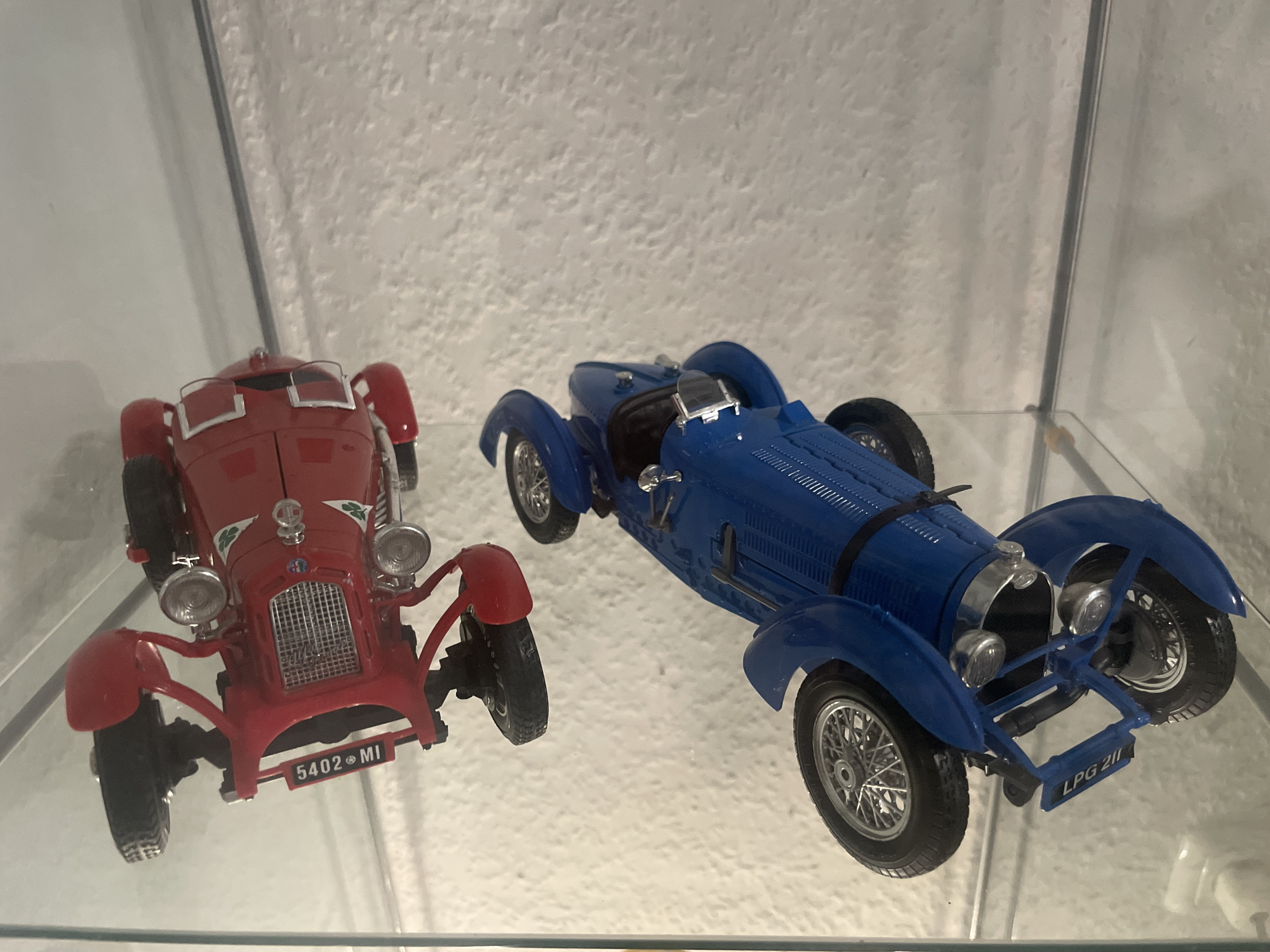 The image shows two vintage toy cars displayed on a glass shelf. The car on the left is red and features a prominent grille, dual headlights, and visible wheels. It has a license plate that reads "5402 MI". The car on the right is blue and has a sleek, aerodynamic design with exposed spoked wheels and a visible windshield. Its license plate reads "LPG 211". The background appears to be a textured wall, possibly inside a display cabinet or on a shelf in a home. The lighting suggests an indoor setting. There are no people or animals present in the image. The scene is clean and orderly, indicating a display of collected items. There is no discernible connection to Iserlohn, Germany.