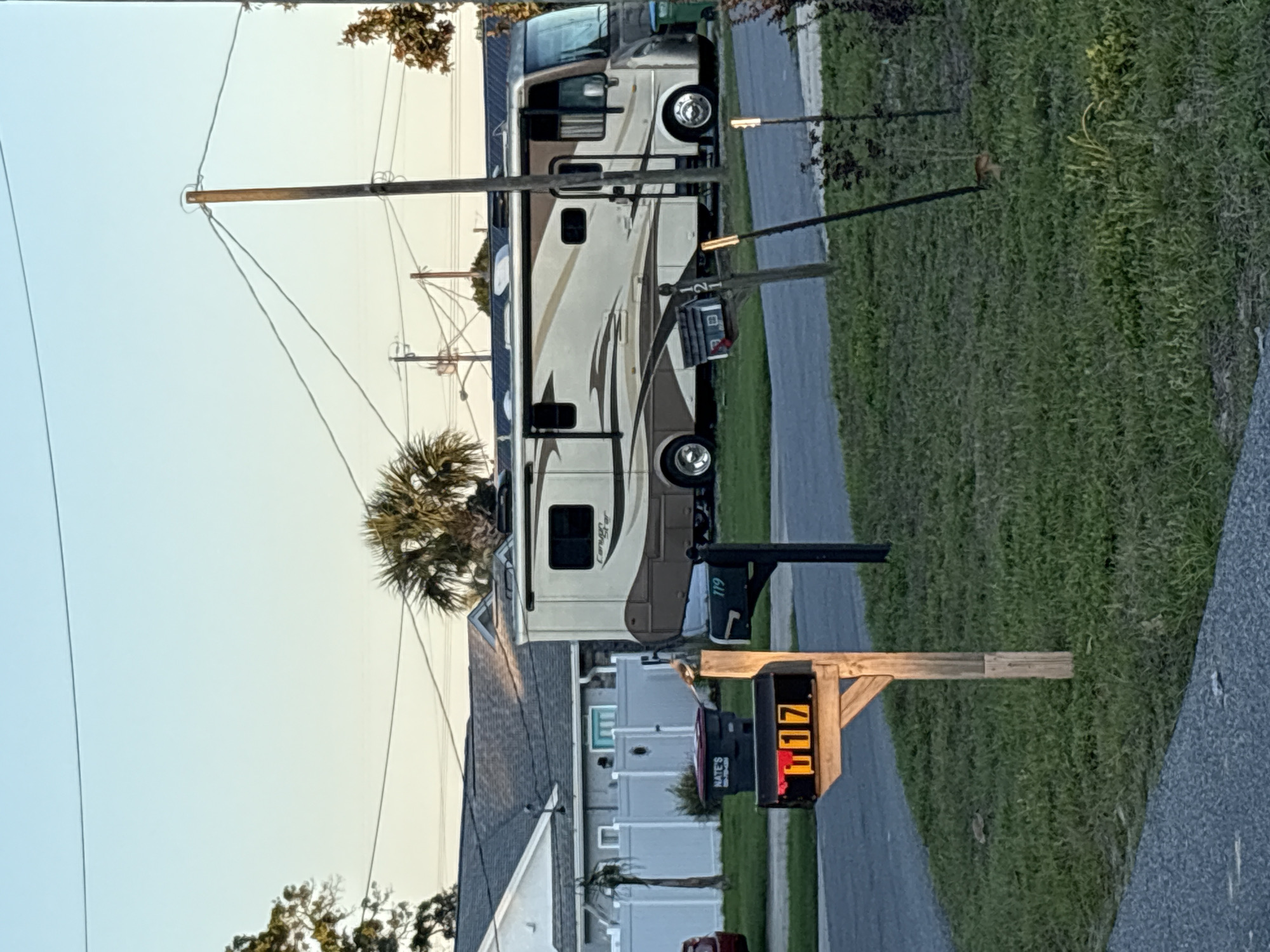 The image depicts a residential street in Panama City Beach, Florida, United States. An RV is parked alongside the road, next to a grassy area. A wooden mailbox post with the number "117" displayed on an illuminated sign is in the foreground. A second black mailbox with a green address plate "119" is further up the street near the RV. A house is visible behind the mailbox, along with a palm tree and utility pole with power lines overhead. The sky is a pale blue, suggesting either early morning or late afternoon.