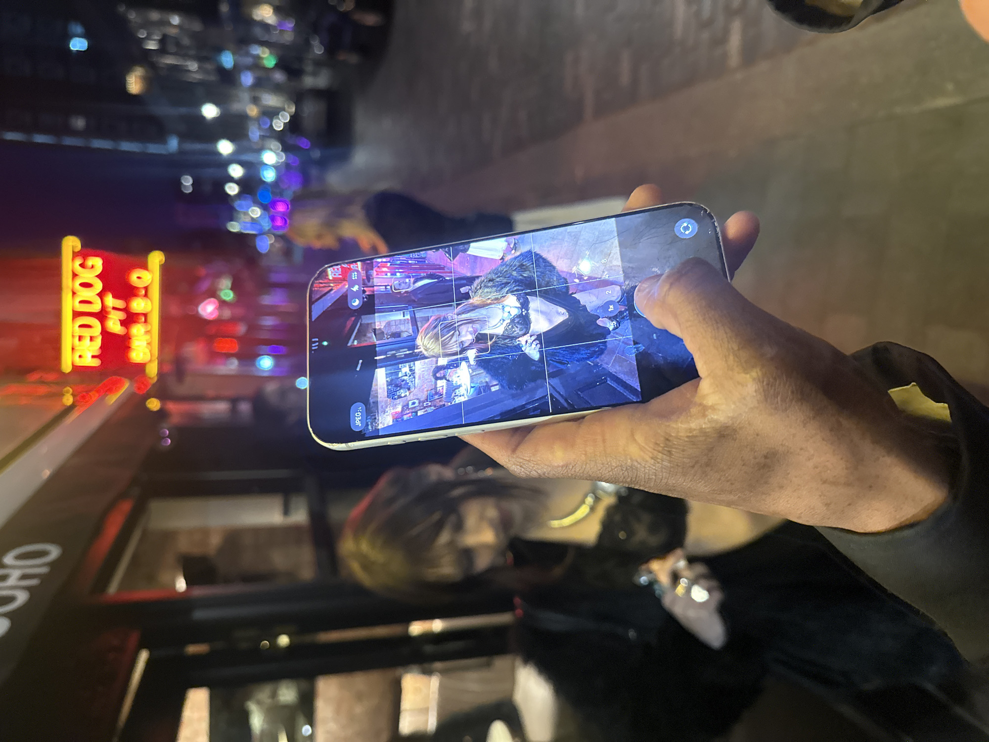The scene depicts a person holding a smartphone, displaying a photo gallery containing an image of a woman wearing a dark top, with a fur collar and blonde hair, potentially outdoors, seated. In the blurred background, a "RED DOG" sign is visible, specifically "RED DOG PIT". "SOHO" is also visible, likely indicating the location in the Soho district of London, United Kingdom. The surrounding environment suggests a nighttime setting on a city street. The person's hand holding the phone is visible in the foreground.