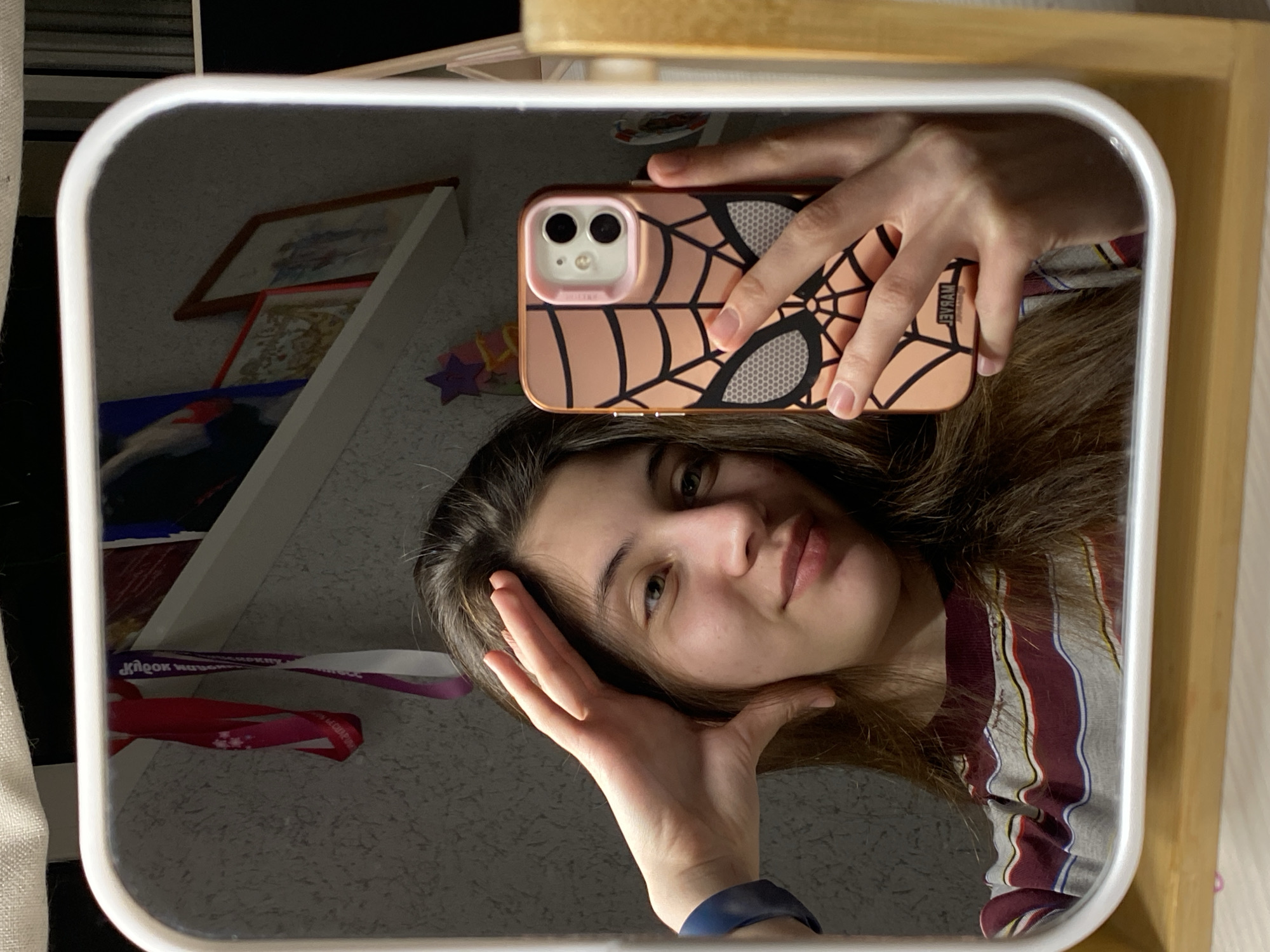 A young woman is captured in a mirror selfie, holding a smartphone with a distinctive peach-colored case featuring a black spiderweb design and textured circular patterns. The reversed text "HYERAM" is visible on the case. Her right hand holds the phone, while her left hand is positioned near her chin and cheek. She has long, dark brown hair and a neutral to slightly smiling expression, looking directly into the mirror. She is wearing a striped shirt with maroon, white, grey, and black vertical lines. A dark blue wristband or watch strap is visible on her left wrist.
The mirror has a white, rounded frame and reflects parts of an indoor setting, likely a personal room. In the background reflection, there are several framed pictures or artworks on a wall, some with wooden frames. A white shelf or object is also reflected. Hanging ribbons or lanyards are visible, one in shades of red and pink, and another in white and purple. The white-purple ribbon has reversed text, partially decipherable as "КІДРОК" (KIDROK) and "WAVEL" or "WAVEY." The overall lighting is somewhat dim in the reflected background, but the mirror frame appears brightly lit. The image is oriented vertically, with the woman's head on the left and her body extending to the right. The location context for this media file is Votkinsk, Russia.