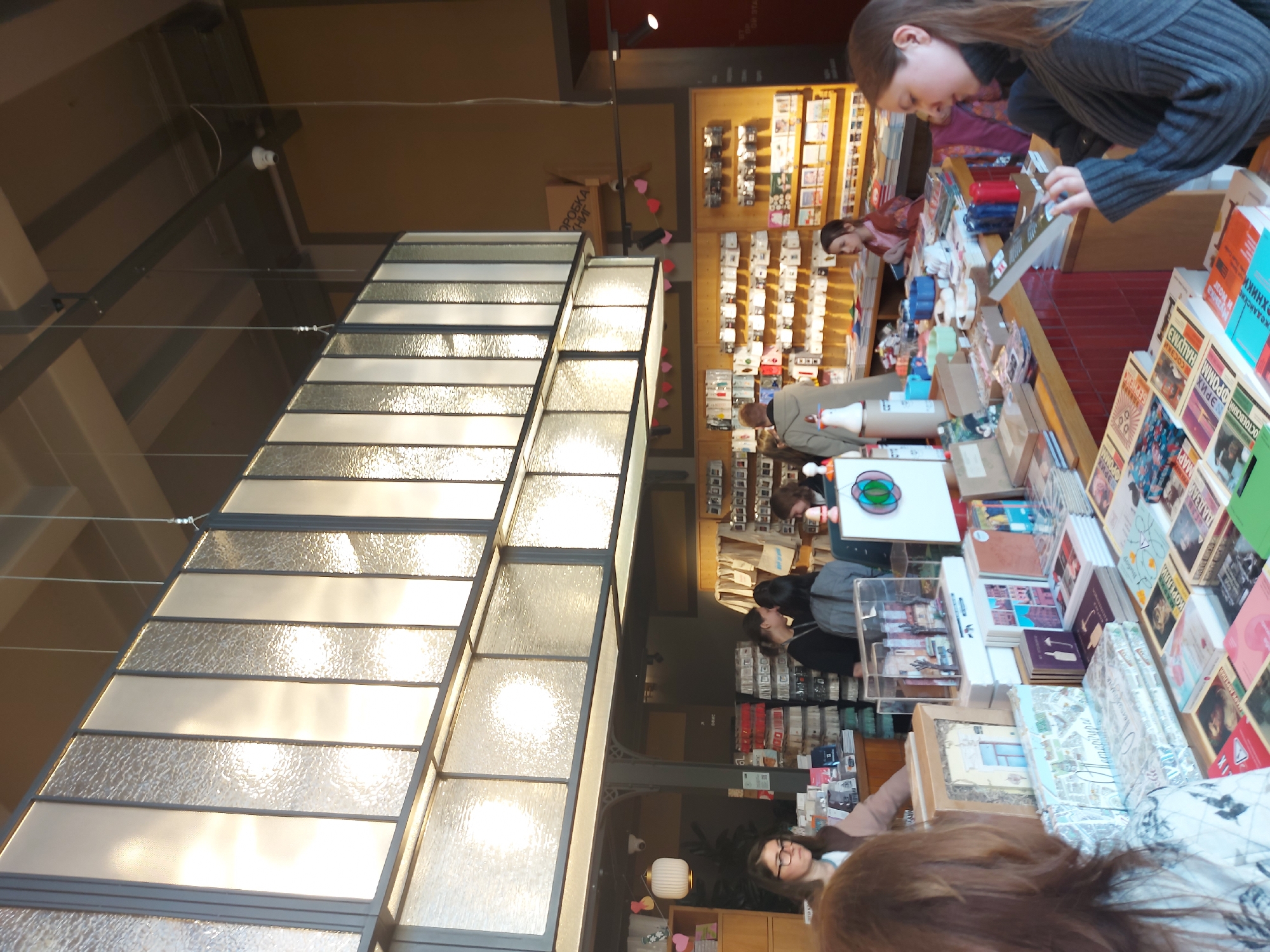 The image depicts an interior scene likely within a bookstore in Saint Petersburg, Russia. There are several people browsing and interacting with merchandise displayed on shelves and countertops. A prominent feature is a series of long, rectangular light fixtures hanging from the ceiling, providing bright illumination. The shelves are filled with books and other goods. A woman is examining items at the counter, while other people are seen looking at the merchandise. The setting appears to be a retail environment catering to the sale of books and other related items.