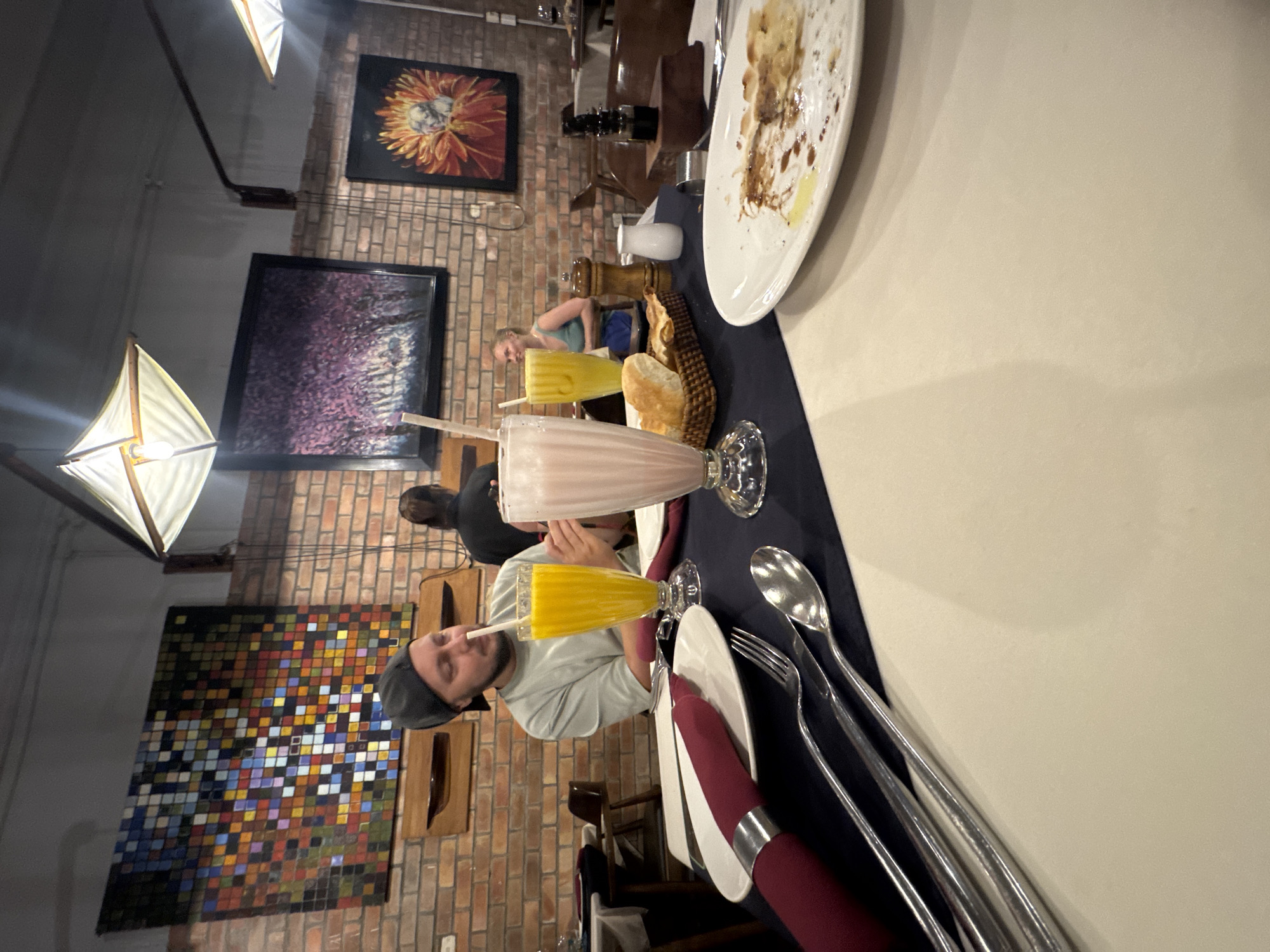 The image captures a scene inside a restaurant, likely located in Hà Nội, Vietnam. A man wearing a grey hat and light blue shirt is seated at a table, drinking from a tall glass with a straw. The table is set with plates, silverware, and cloth napkins. Two drinks, a yellow one and a pink one, are placed on the table. Behind the man are two other patrons. The brick wall behind them is adorned with several framed art pieces, including a mosaic, a framed portrait of a sunflower, and a framed piece showcasing a dark abstract pattern. Overhead, modern, geometric light fixtures illuminate the scene.
