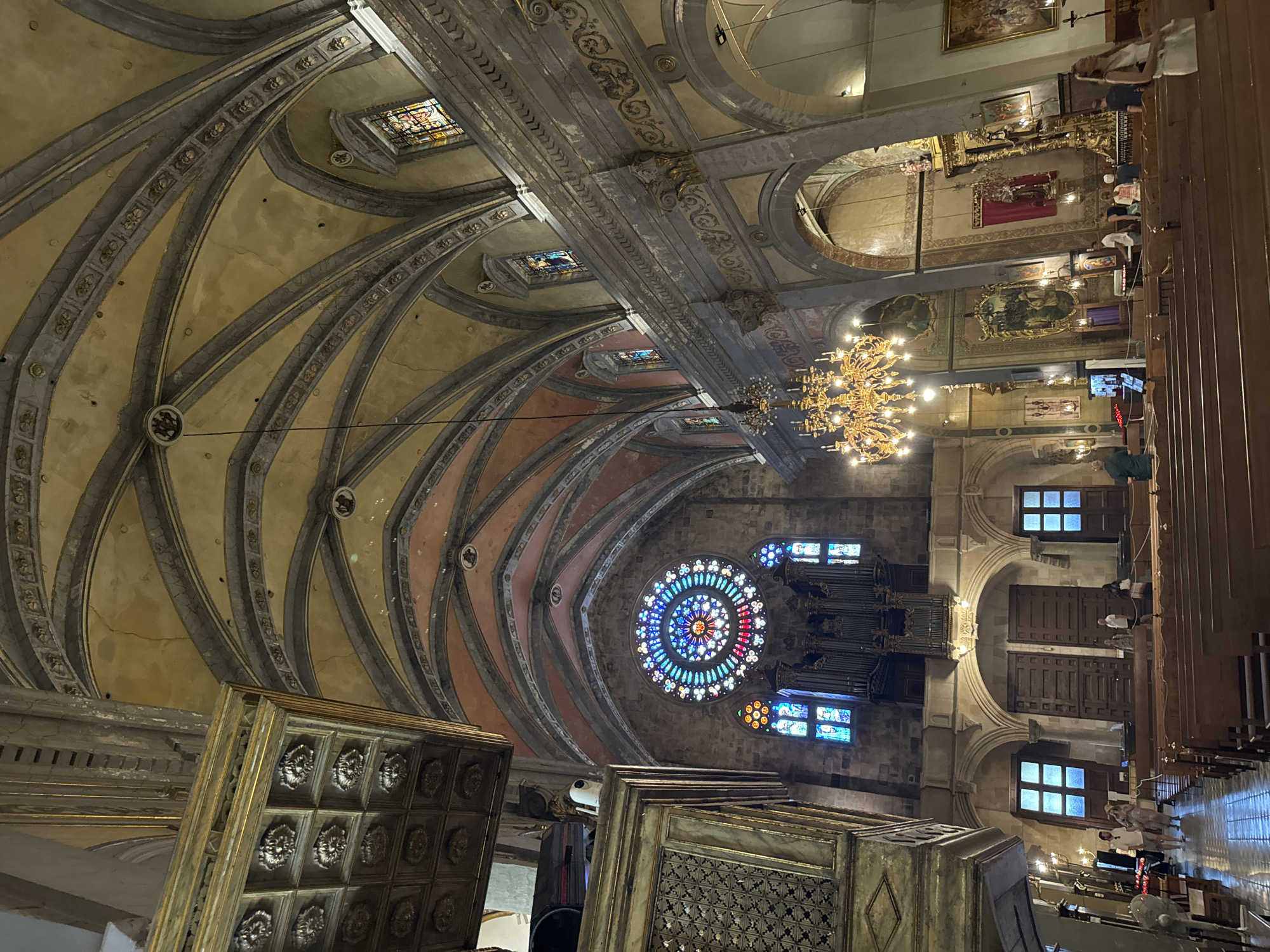 The image depicts the interior of a grand church with a high, arched ceiling. Sunlight streams through a large, circular stained-glass window, known as a rose window, which is centrally located above a pipe organ. Smaller stained-glass windows are also visible on either side. A large, ornate golden chandelier hangs in the middle of the nave, casting a warm glow. Wooden pews are arranged in rows, and a few individuals are scattered throughout the church, some standing and others seated. The architecture is a blend of Gothic and Baroque styles, with intricate detailing on the walls and ceiling. This church is located in the city of Sóller, Spain, and is a significant historical and religious landmark.