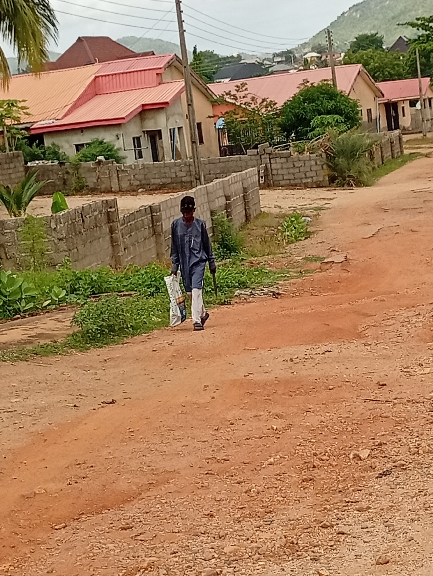 In Adankolo, Nigeria, an individual is walking on a dirt road. The person is dressed in dark, traditional attire and carrying a bag. Along the road are brick fences and houses with red roofs. A mountain is visible in the distance. Overhead electrical lines are present. The ground is reddish-brown dirt with loose gravel. Patches of green vegetation are interspersed along the road.