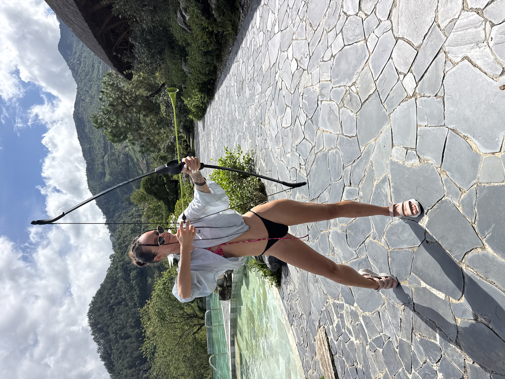 This image captures a woman in a dynamic pose, holding a black bow, in an outdoor setting identified as Púng Luông, Vietnam. She stands on a grey stone-paved path, with her body angled and leaning forward, as if preparing to shoot an arrow. The woman is dressed in a black bikini bottom, a light-colored long-sleeved shirt worn open over a pink patterned bikini top, and light pink sandals. Sunglasses are pushed up onto her head.
To her left, a swimming pool with shimmering blue water is partially visible, bordered by the same stone paving. The background features a breathtaking vista of towering, lush green mountains covered in dense vegetation, stretching towards a bright blue sky dotted with white clouds. The strong sunlight casts distinct shadows on the ground, indicating it is daytime, likely late morning or afternoon. A light green, funnel-shaped object on a stick protrudes from behind some foliage near the path. A glimpse of a wooden architectural structure is visible at the top left, suggesting a resort or lodge integrated into the natural landscape. The scene evokes a sense of adventure and leisure amidst a picturesque mountainous environment. No visible text is present in the image.