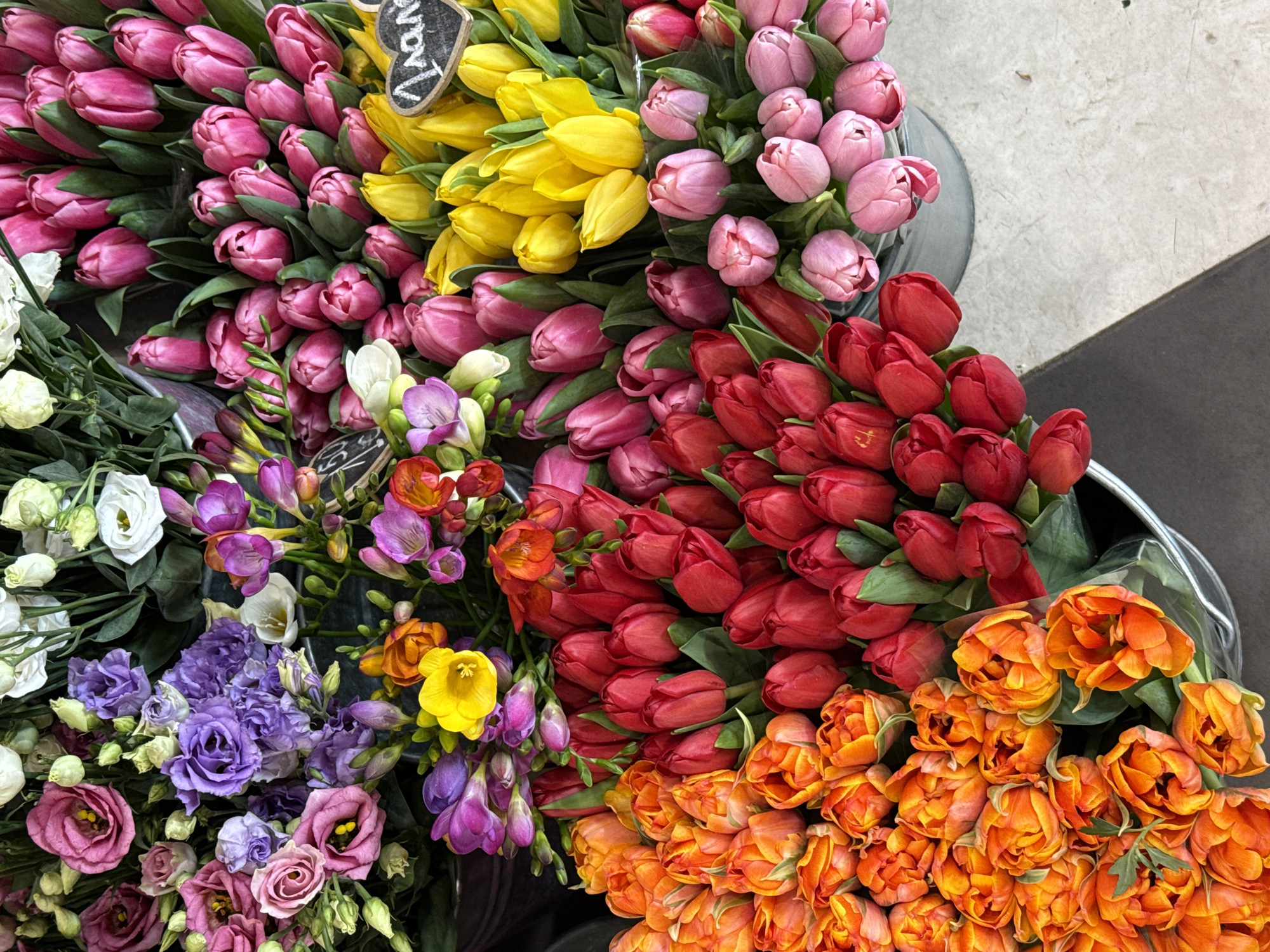 The image shows an assortment of flowers arranged close together, possibly for sale. Bunches of tulips dominate the composition, with colors ranging from pink and red to yellow and orange. Other flowers, including freesia and lisianthus in white, purple, and pink, are also present. The setting could be a flower market or a flower shop, possibly located in the city of Plovdiv, Bulgaria.