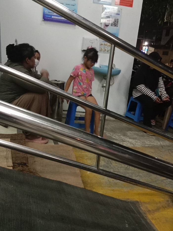 The image depicts an indoor public waiting area or corridor in Gladaksari, Indonesia, under artificial lighting, with a darker exterior visible on the right. The foreground is dominated by two parallel stainless steel handrails, indicating a ramp or steps.
On the left side of the frame, two adult females are positioned near a silver cylindrical trash can. One female faces away from the camera, wearing a dark top, light brown pants, and pink slip-on sandals. The other female, partially obscured behind the first, wears a white face mask and a light-colored top, appearing to be in conversation.
In the center, a young child, presumably female, with dark hair and glasses, wears a pink t-shirt with a colorful pattern and dark shorts. The child is barefoot and seated on a blue plastic stool, looking downwards and slightly to the right.
Behind the child, a light blue wall-mounted sink with a faucet is affixed to a white wall. Below it, a white dispenser is mounted, and above are several signs, including a blue informational sign and a red-and-white sign with a QR code labeled "PENDAFTARAN ONLINE" (Online Registration).
To the far right, partially obscured by the railing and ambient darkness, an adult wearing a black hijab and a black-and-white striped long-sleeved shirt is seated on a second blue plastic stool.
The floor in the foreground transitions from a dark, ribbed anti-slip mat to a light brown textured surface, and then to a lighter concrete floor marked with a yellow painted stripe. The overall ambient light suggests evening or nighttime conditions.