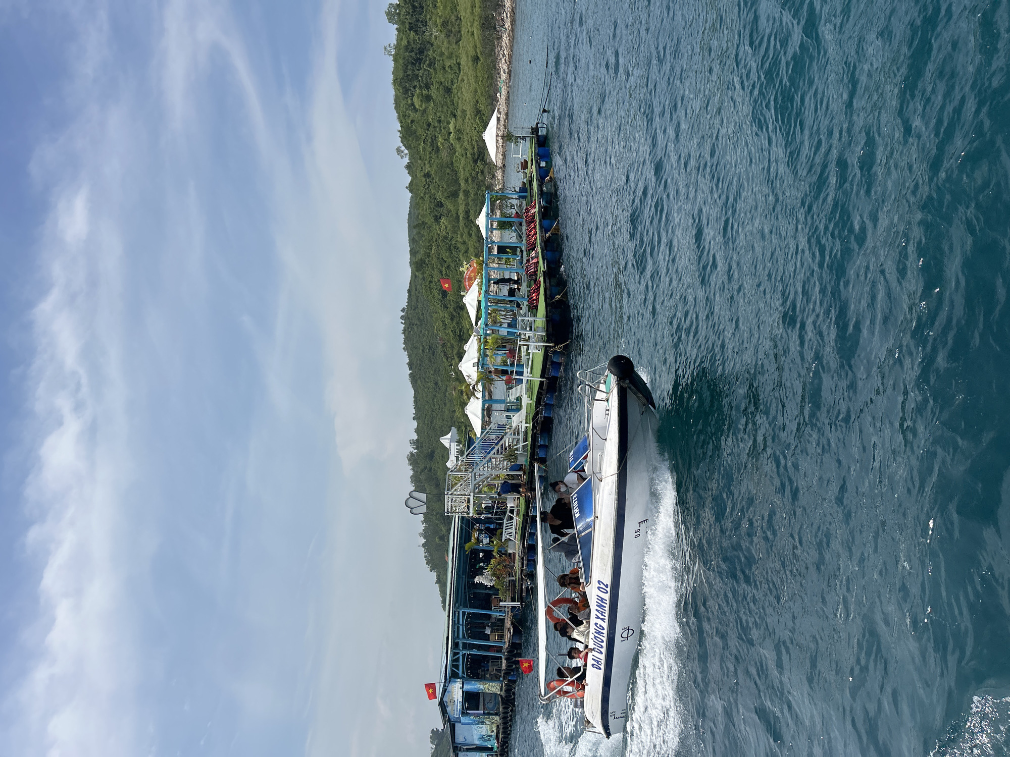 An eye-level medium shot captures a speed boat with several passengers moving across turquoise water in Nha Trang, Vietnam. The boat, labeled "DAI DUONG XANH 02", is white with a blue stripe and a white canopy. A Vietnamese flag flies from the boat's stern. In the background, a floating platform with white umbrellas, lounge chairs, and life vests is visible. Beyond the platform, a lush green island with a small white structure on its peak rises from the sea, with more smaller islands and the mainland in the distance. The sky is a clear blue with scattered white clouds. The water is choppy, indicating the boat's movement. The overall scene suggests a recreational outing in a coastal area.