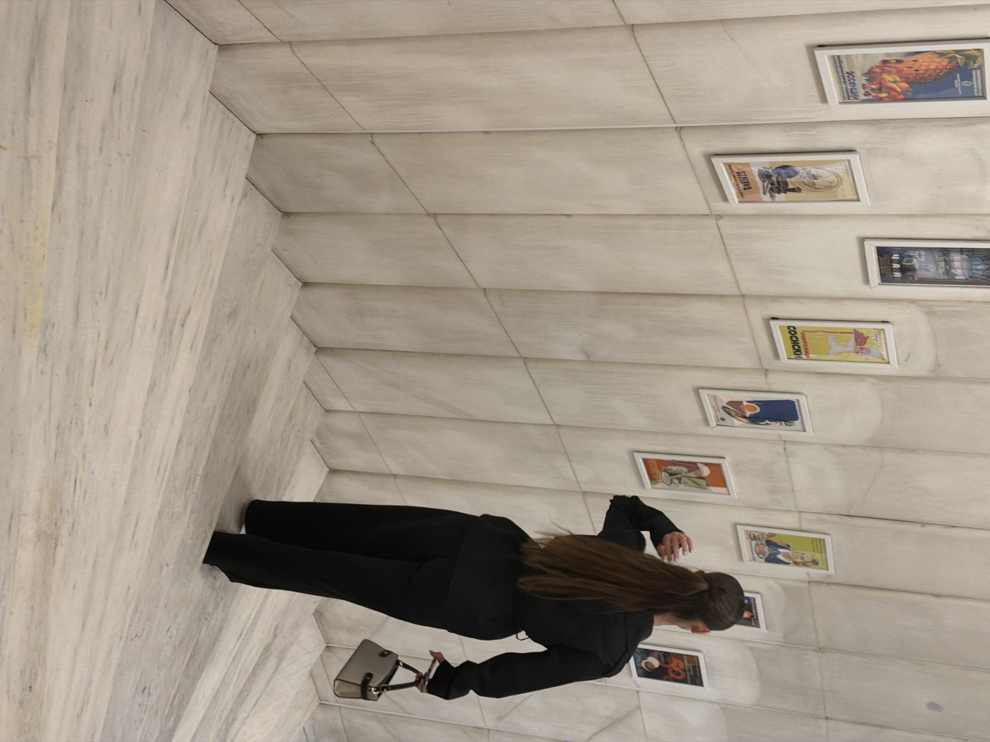 A woman in black clothing stands in what appears to be an immersive art installation in Sofia, Bulgaria. The installation is designed to look like an Escher-esque space, with walls, floor, and ceiling tiled in a pale marble pattern, creating a disorienting perspective. The woman is facing the wall, looking at several framed posters that are mounted in a vertical line. She is holding a gray purse.