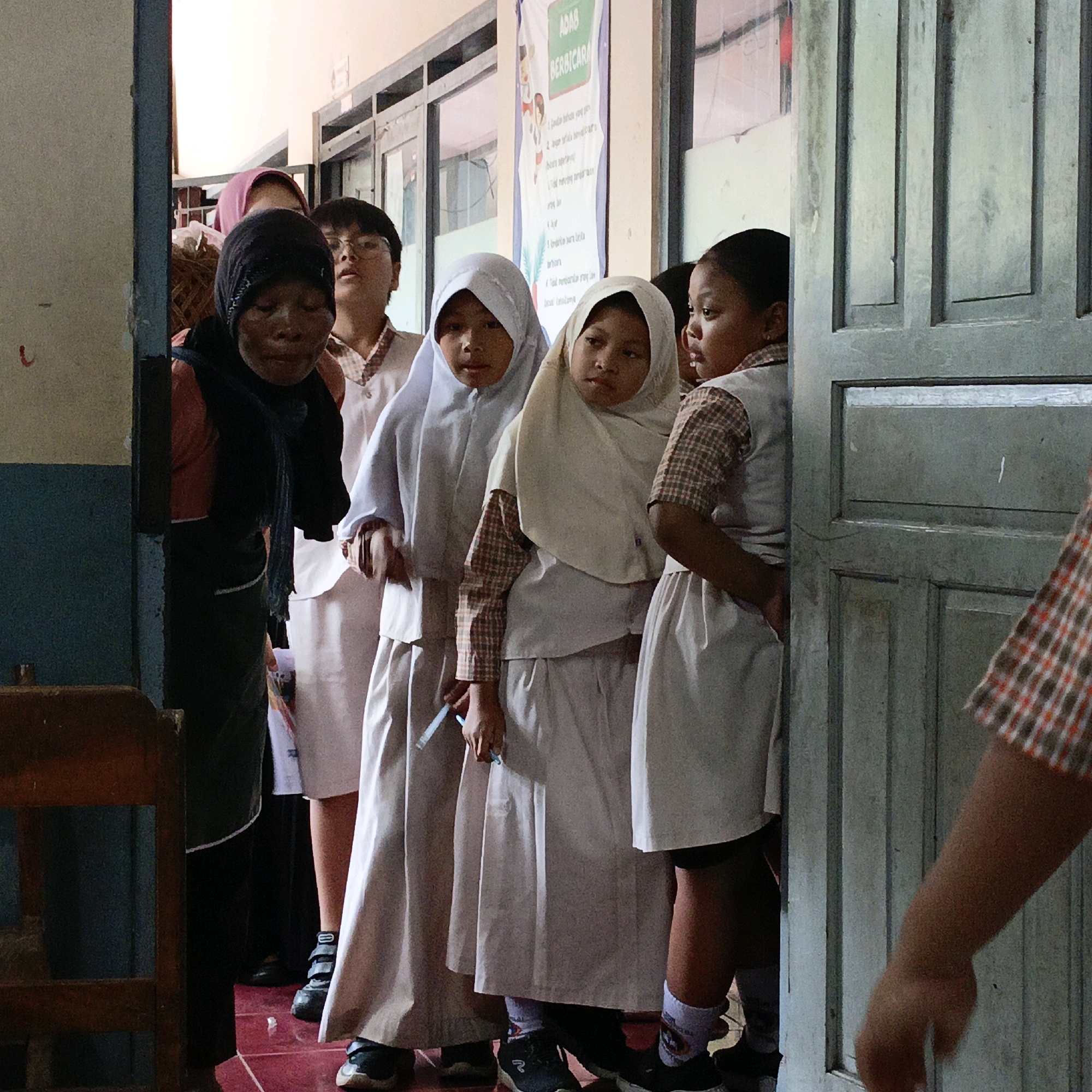 The image depicts a group of school children in uniform standing in a doorway in Salatiga, Indonesia. The children are wearing white hijabs and uniforms with plaid accents on their shirts. Some of the children are holding pencils. A poster with text in Indonesian is visible on the wall behind them. The doorway is made of weathered blue wood. The flooring is a dark reddish-brown tile. The scene appears to be a school hallway.