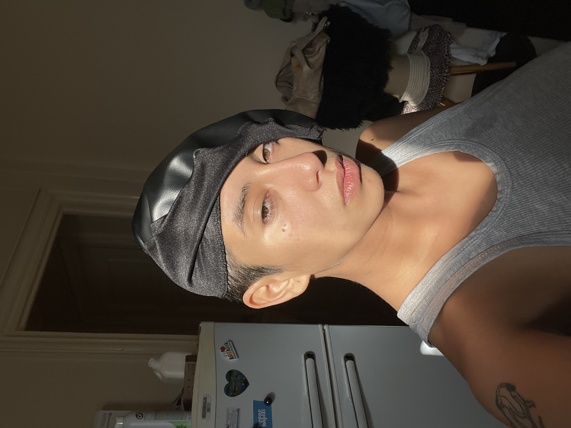 A person with fair skin, wearing a black head covering and a gray tank top, is depicted in a medium shot. The person is positioned in front of a light gray refrigerator with several magnets attached. The setting appears to be indoors, possibly a kitchen or apartment. The lighting is strong, with a direct ray of sunshine illuminating the person's face and upper body. No specific location information related to Buenos Aires, Argentina is evident in the scene.