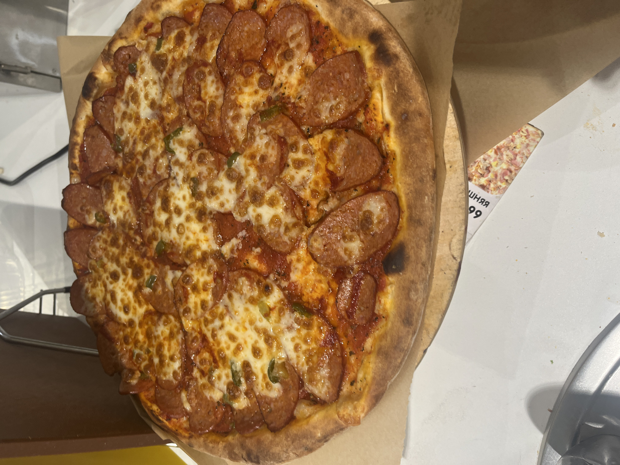A close-up overhead shot shows a pepperoni pizza on a circular wooden board, partially obscured by a brown paper bag. The pizza features a golden-brown crust, melted mozzarella cheese, tomato sauce, and numerous circular slices of pepperoni. Small green pieces, likely peppers, are scattered across the surface. A partially visible receipt with the number "66" and Cyrillic text is tucked beneath the pizza board. The background consists of a white surface, a brown wooden object on the left, and metal shelving in the upper right corner, suggesting an indoor setting such as a restaurant kitchen or a delivery service hub. There is no indication of the city of Kaliningrad, Russia, in the image.