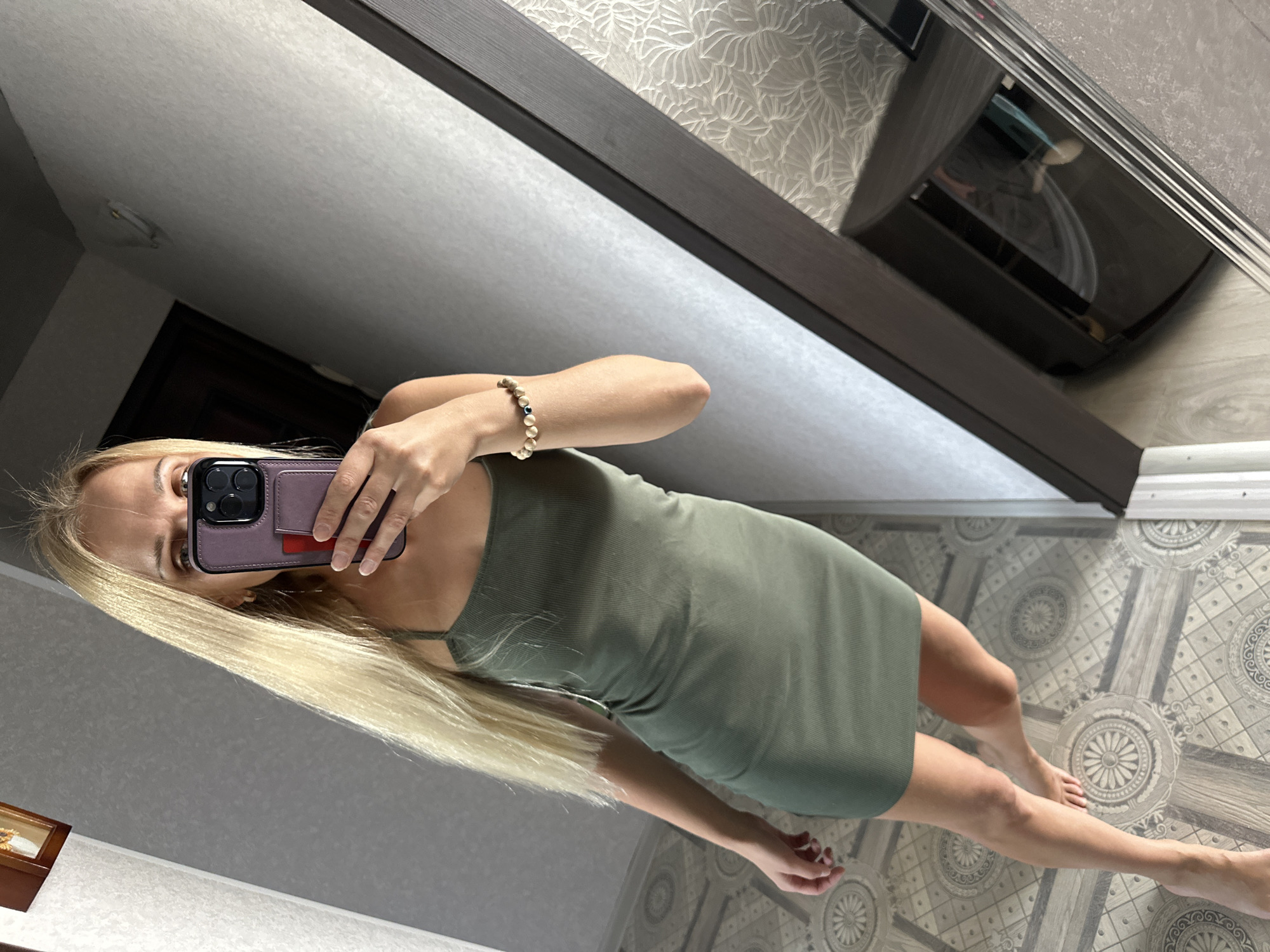 A woman with long blonde hair is taking a mirror selfie in what appears to be a domestic setting. She is wearing a form-fitting olive green ribbed dress and a beaded bracelet on her left wrist. Her right hand is holding a dark purple phone case with a phone inside. The floor has a patterned tile design. In the reflection of a mirror to the right, part of a dark wooden cabinet and a wall with textured wallpaper are visible. The lighting suggests it is daytime. The scene is not identifiable as being in the city of Energetik, Russia.