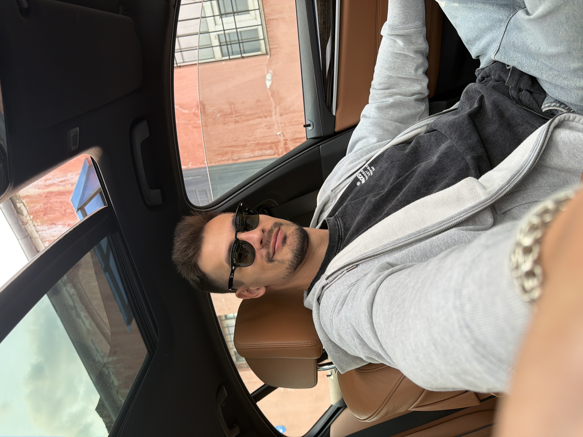 The image shows a man sitting in the driver's seat of a car. He is wearing sunglasses, a grey zip-up hoodie over a dark t-shirt, and blue jeans. The interior of the car has brown leather seats and a sunroof. Outside the car, a building with orange-pink walls and windows is visible. The image does not provide any specific information or visual cues that would allow for the identification of the location as Krasnoyarsk, Russia.
