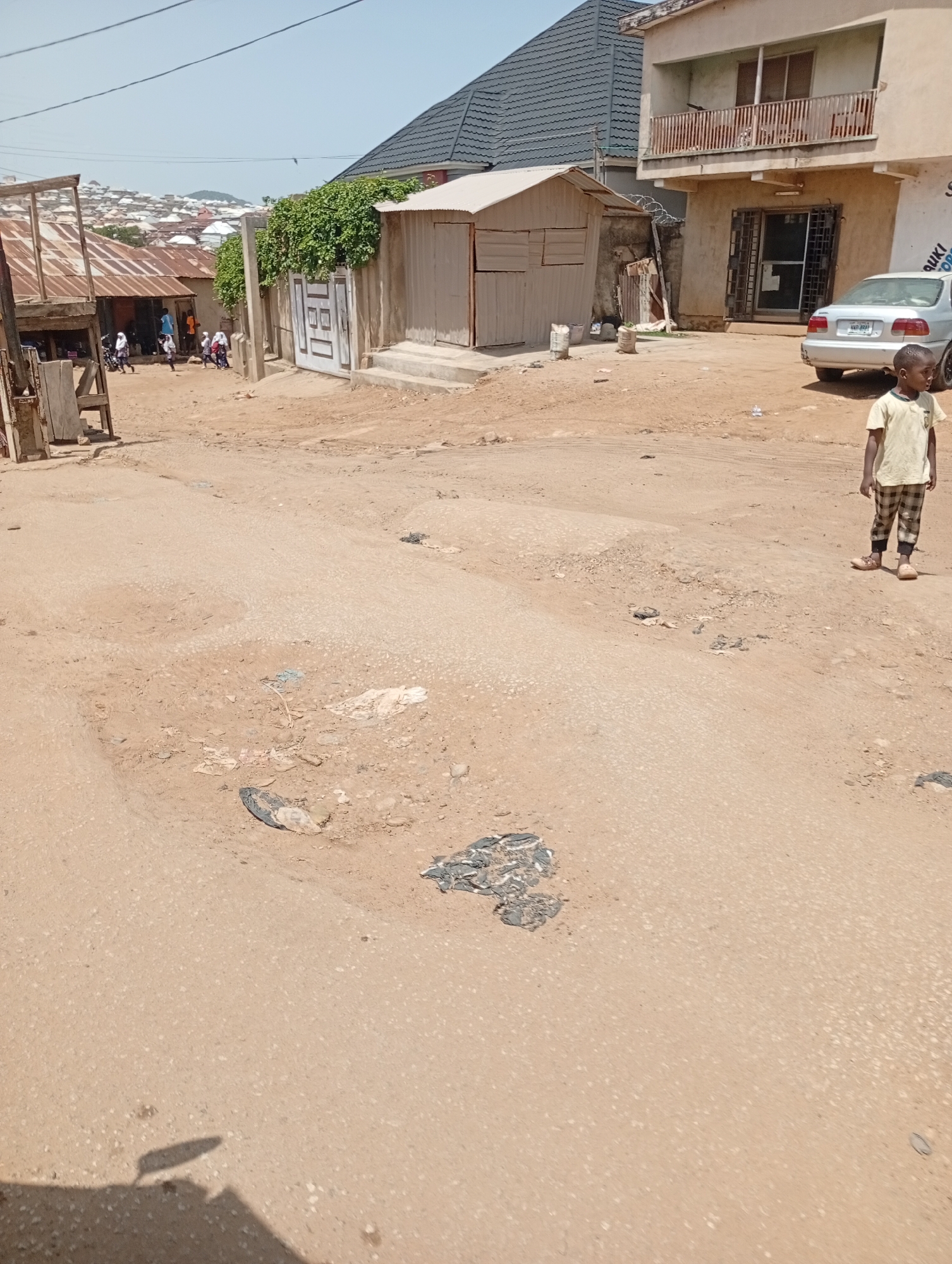 This image depicts a street scene in Jos, Nigeria, characterized by a dirt road with visible potholes and debris. In the foreground, a young boy stands facing right, wearing a light yellow t-shirt and checkered pants. Behind him, a silver car is parked near a building with a balcony. To the left, a small, corrugated metal structure stands adjacent to a larger building with a dark, tiled roof. In the background, a cluster of buildings with tin roofs stretches across a hillside, suggesting a densely populated area. Several people, some appearing to be children, are visible in the middle ground near the structures. The overall environment appears to be an urban setting with signs of infrastructure development and everyday life.