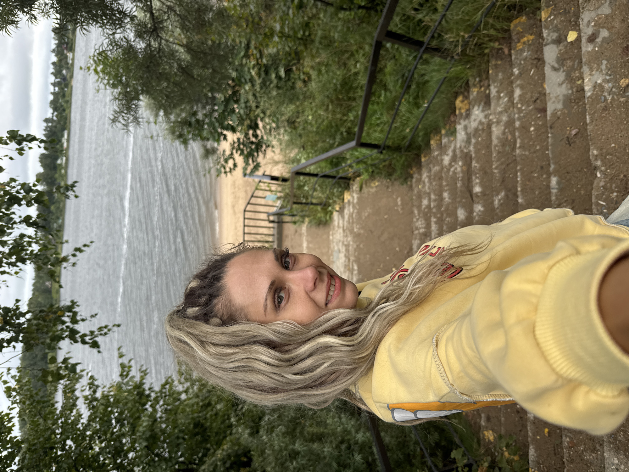 A young woman with blonde dreadlocks pulled into a high ponytail is taking a selfie on a set of concrete stairs that lead down to a sandy beach and a body of water. She is wearing a yellow sweatshirt. The beach and water are in the background, with trees and foliage on either side of the stairs. The sky is overcast. The location is identified as Rusanovo, Russia.