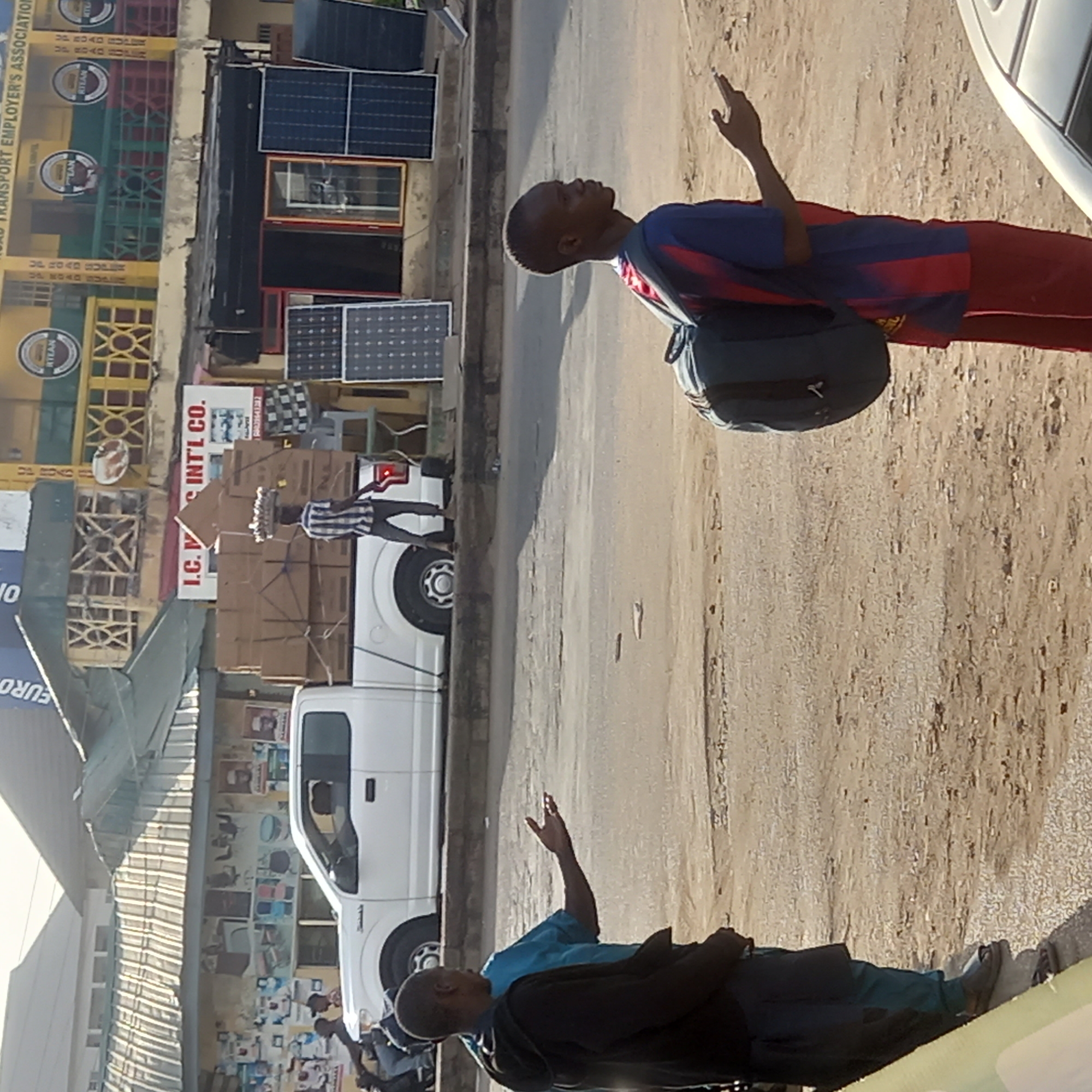 The photograph appears to be taken on a street in Sardauna, Nigeria. The background features buildings and signage, including "ROAD TRANSPORT EMPLOYER'S ASSOCIATION," "RTEAN," and "I.C. INT'L CO." Solar panels are also visible on the facade of one of the buildings. A white pickup truck is parked in the street. Two men are standing on the side of the road. One man is wearing a blue and red striped shirt with red pants and a backpack. The second man is wearing a light blue shirt and darker pants and jacket. Both men are gesturing with their hands in the air. The street is paved and appears to be dusty. Other people can be seen in the background.