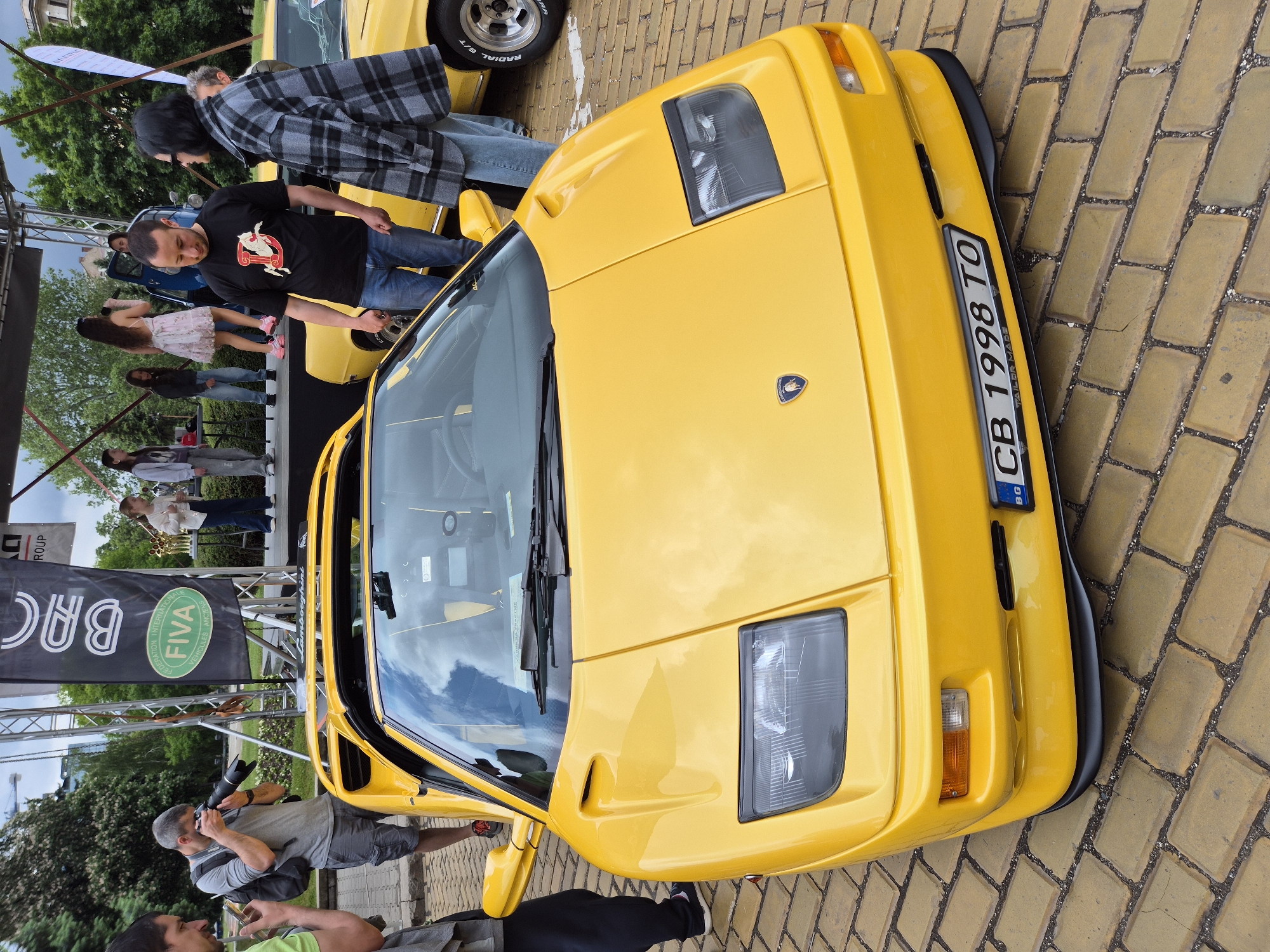 The image shows a yellow Lamborghini Diablo parked on a cobblestone street. The license plate reads "CB 1998 TO", which is a Bulgarian license plate. People are standing around the car, some appearing to be admiring it or taking pictures. There is a banner in the background with the logos of "BAC" and "FIVA" on it. The scene appears to be at an outdoor car show or event, likely in Sofia, Bulgaria.