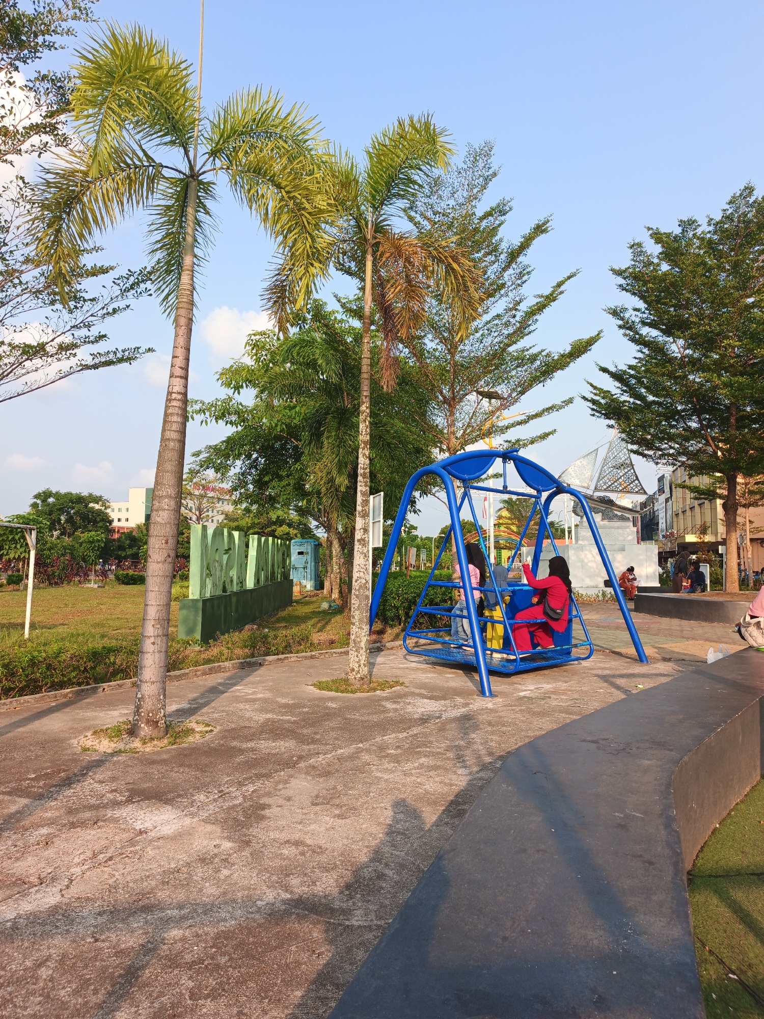 This image depicts a park scene in Batam, Indonesia. Two palm trees are prominent in the foreground. To the right, a blue metal swing set is occupied by at least two individuals, one wearing a red outfit. In the background, there's a lawn with a green sculpture or sign. Further back, buildings and trees are visible, suggesting an urban park setting. The sky is clear and blue. The ground is paved with concrete, and there is a dark, curved wall or seating area on the right side of the frame. The lighting suggests it is late afternoon or early evening.