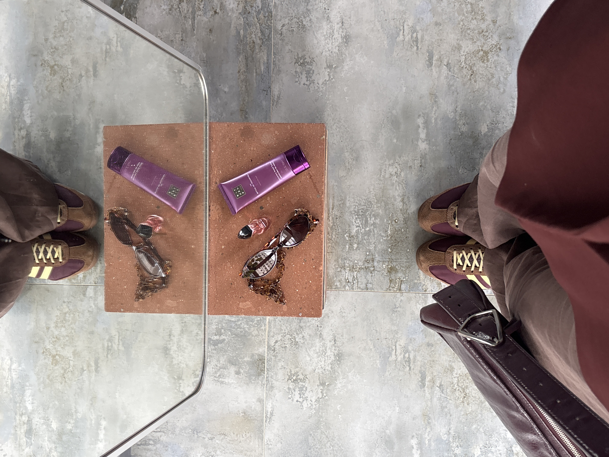The image shows a top-down view of a tiled floor with a small terracotta-colored table in the center. On the table are two purple tubes of cream, a small perfume bottle, and sunglasses with a chain. To the left of the table is a mirror reflecting the items. In the bottom corners of the image, parts of two people are visible. The person on the left has on brown sneakers with yellow stripes, while the person on the right has on brown shoes and is carrying a brown leather purse. The environment appears to be indoors on a tiled floor. The location in the city of Yerevan, Armenia cannot be determined from the given image.