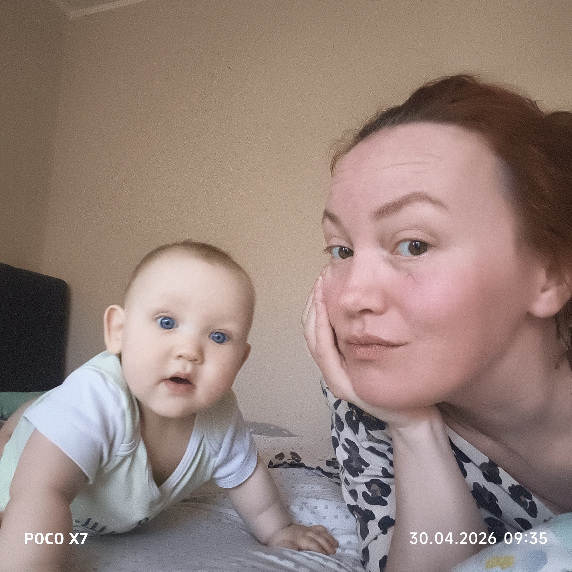 This media file captures a close-up portrait of a woman and a baby, likely taken indoors in Chaikovsky, Russia. The setting appears to be a bed with light-colored bedding, against a plain light brown wall.
On the right side of the frame, a woman with reddish-brown hair, pulled back, rests her chin on her hand, looking towards the camera. She wears a black and white leopard print top. Her expression is calm and gentle.
To her left, an infant with light hair and striking blue eyes is positioned on its hands and knees, looking directly at the camera with an open mouth, conveying curiosity. The baby wears a light green and white onesie. A dark piece of furniture, possibly a headboard, is partially visible in the upper left background.
The image is brightly lit, suggesting natural daylight. Visible text in the bottom left corner reads "POCO X7", indicating the device used for the photograph. A timestamp in the bottom right corner shows "30.04.2026 09:35". The overall scene depicts an intimate, loving interaction between a mother and child.