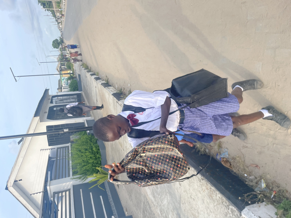 An outdoor scene in Mosafejaw, Nigeria, depicts a group of students on a dirt and paved roadside. In the foreground, a male student, appearing to be of elementary school age, is dressed in a white short-sleeved collared shirt with a red bow tie, blue and white checkered shorts, and dark sneakers with white socks. He carries a black shoulder bag on his left side and holds a patterned, glossy black and gold drawstring backpack in both hands, looking down at it. His expression is focused downwards. Behind him, another male student is partially visible, wearing blue shorts and dark shoes.
To the student's left, a paved sidewalk with a black and white striped curb runs alongside the scene. Beyond the sidewalk, a white and gray building with a gated fence and security wire is visible. Green shrubs grow in front of the building. Further down the sidewalk, another student wearing a gray uniform and a black backpack walks away from the camera.
In the mid-ground and background, a wide, unpaved road stretches into the distance. Several other students, predominantly female, wearing various school uniforms and carrying bags, walk along the right side of the road away from the camera. Utility poles with streetlights line the road. The sky is clear with scattered clouds. The overall lighting suggests daytime.