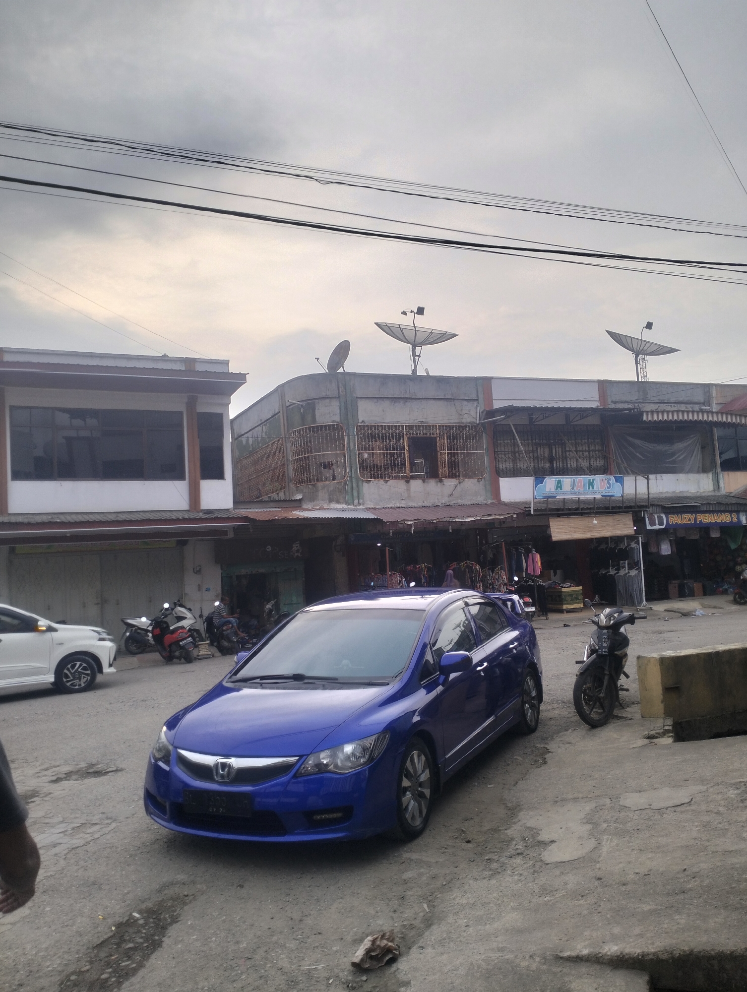 A blue sedan is parked on a paved surface. The car has its headlights on and its front license plate is visible. A black motorcycle is parked to the right of the sedan. In the background, there are buildings with satellite dishes on the roofs. Power lines are strung across the sky. The environment appears to be an urban setting in Keudegeudong, Indonesia.