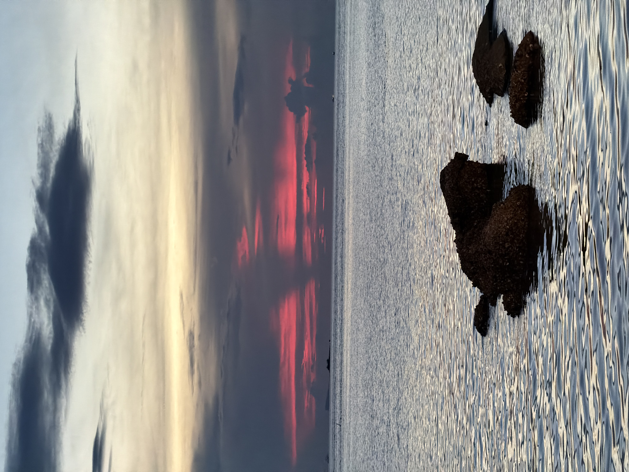 The image depicts a serene seascape with a dramatic sunset over the ocean. The sky is a canvas of colors, with streaks of fiery red and pink contrasting with softer hues of yellow and grey, suggesting the sun is setting or has just set. Dark, wispy clouds add texture to the sky, with one particularly sharp cloud formation resembling a pointed spike on the left side.
The ocean occupies the right side of the frame, its surface rippled by gentle waves that reflect the sky's light, creating a shimmering effect. Several dark, rough-textured rocks are partially submerged in the water, anchoring the scene and providing a strong visual element. Their dark silhouettes stand out against the lighter, reflective water. In the distance, a faint outline of land or a larger landmass can be seen on the horizon, with a few small, dark shapes that could be boats or structures.
The overall mood of the image is peaceful and contemplative, evoking the beauty of nature at dusk. The play of light and shadow, the vibrant colors of the sky, and the tranquil water create a sense of calm. The location context provided, Baan Bo Phut, Thailand, suggests a tropical or subtropical coastal environment. There is no visible text in the image.