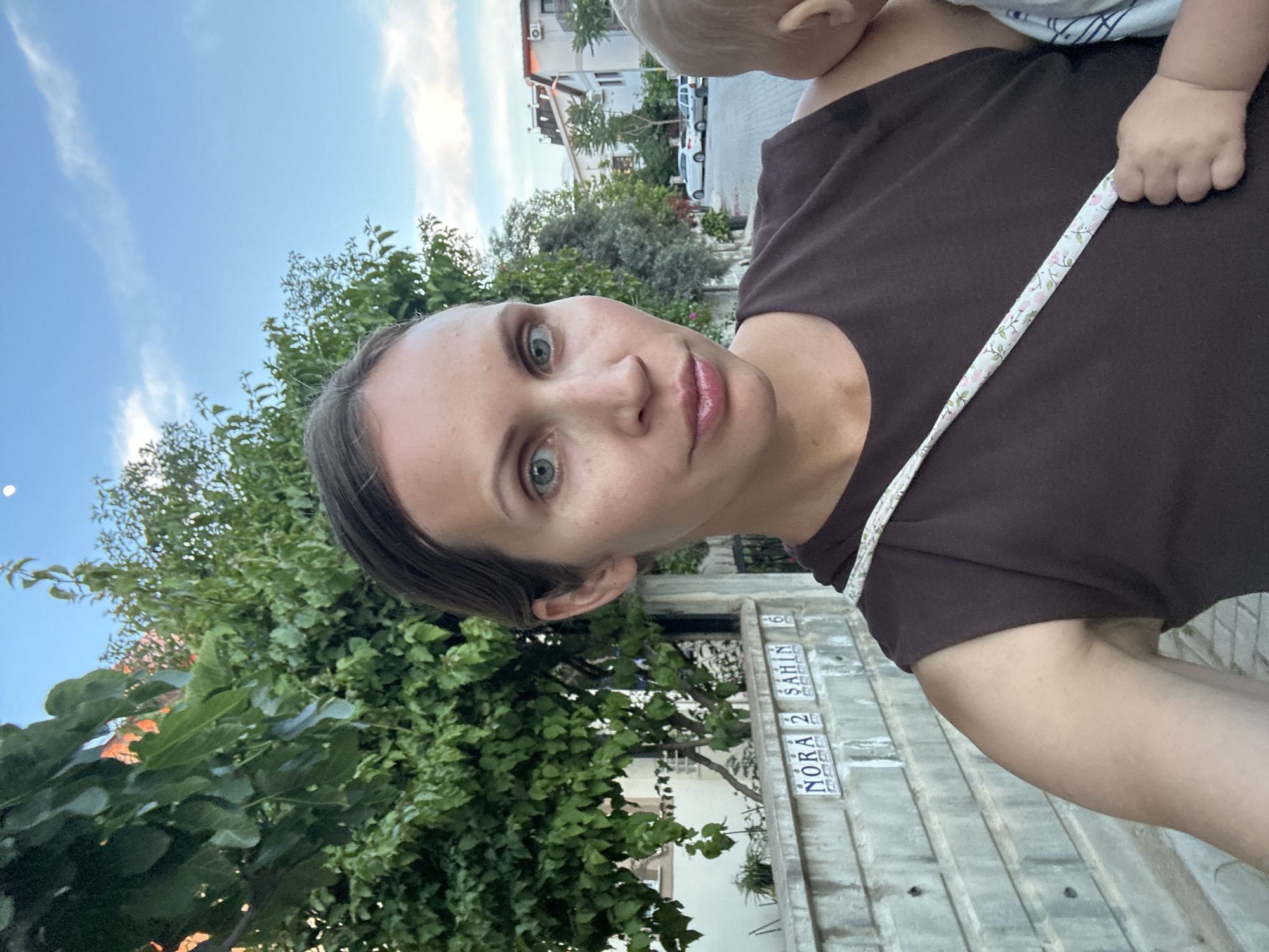 A woman holds a baby on her shoulder, with a tree and a building behind her. The building has a sign that says "NORA 2 SAHIN", placing the location in Fethiye, Turkey.