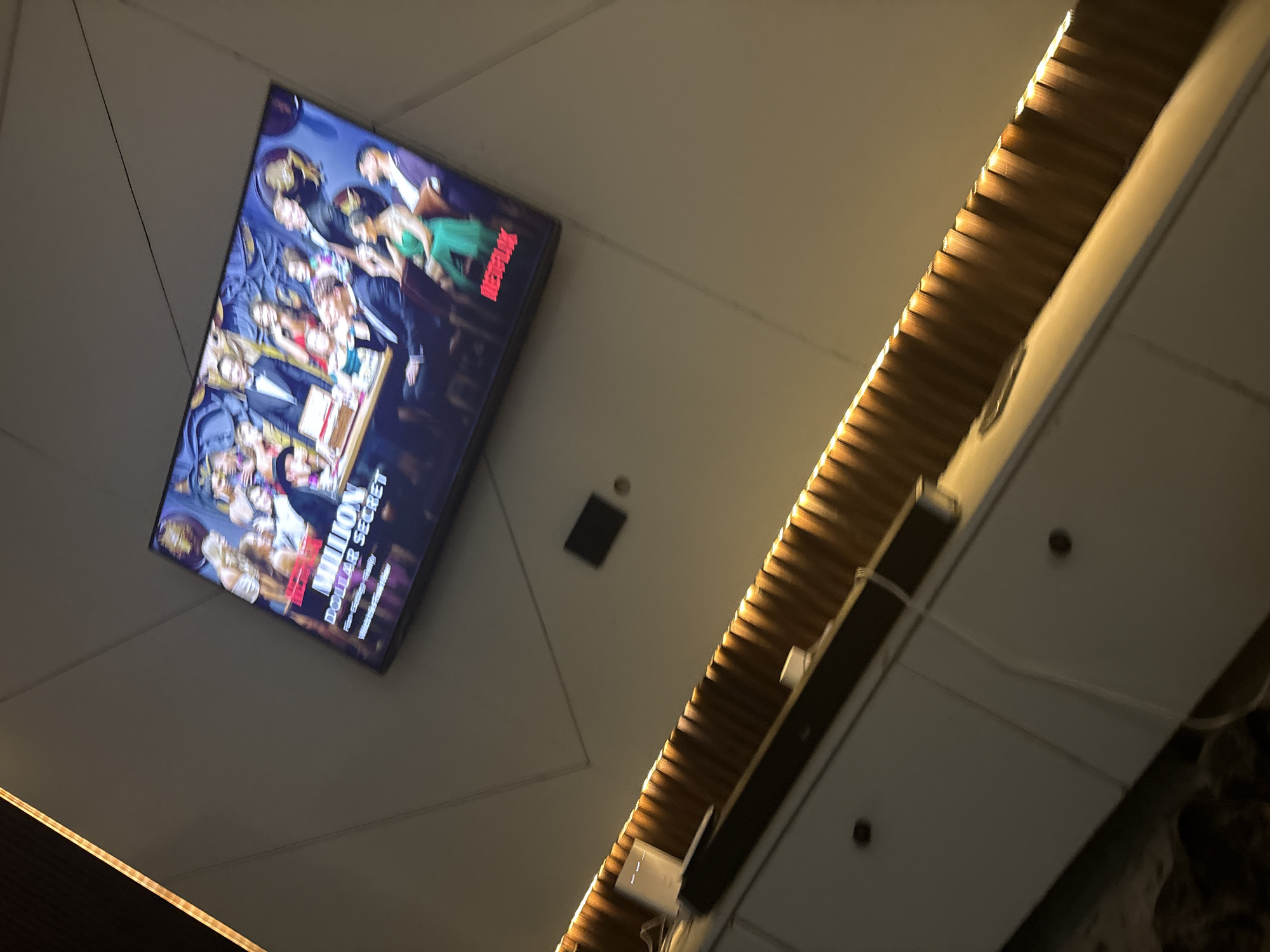 The image shows a television screen displaying the Netflix show "Million Dollar Secret" mounted on a ceiling. A recessed lighting fixture runs diagonally across the ceiling. There is no evidence to suggest that this image was taken in Iddo Tudunwada, Nigeria.