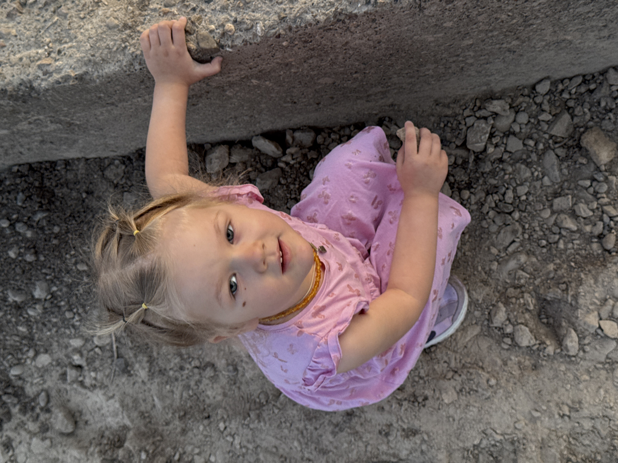 A young child in a pink dress and yellow beaded necklace stands on a rocky surface, grasping a concrete wall with one hand and holding a small rock in the other. The scene appears to be outdoors, with the child positioned near a concrete structure and surrounded by gravel and rocks. The perspective is from directly above, looking down at the child. The location is Горный, Russia.