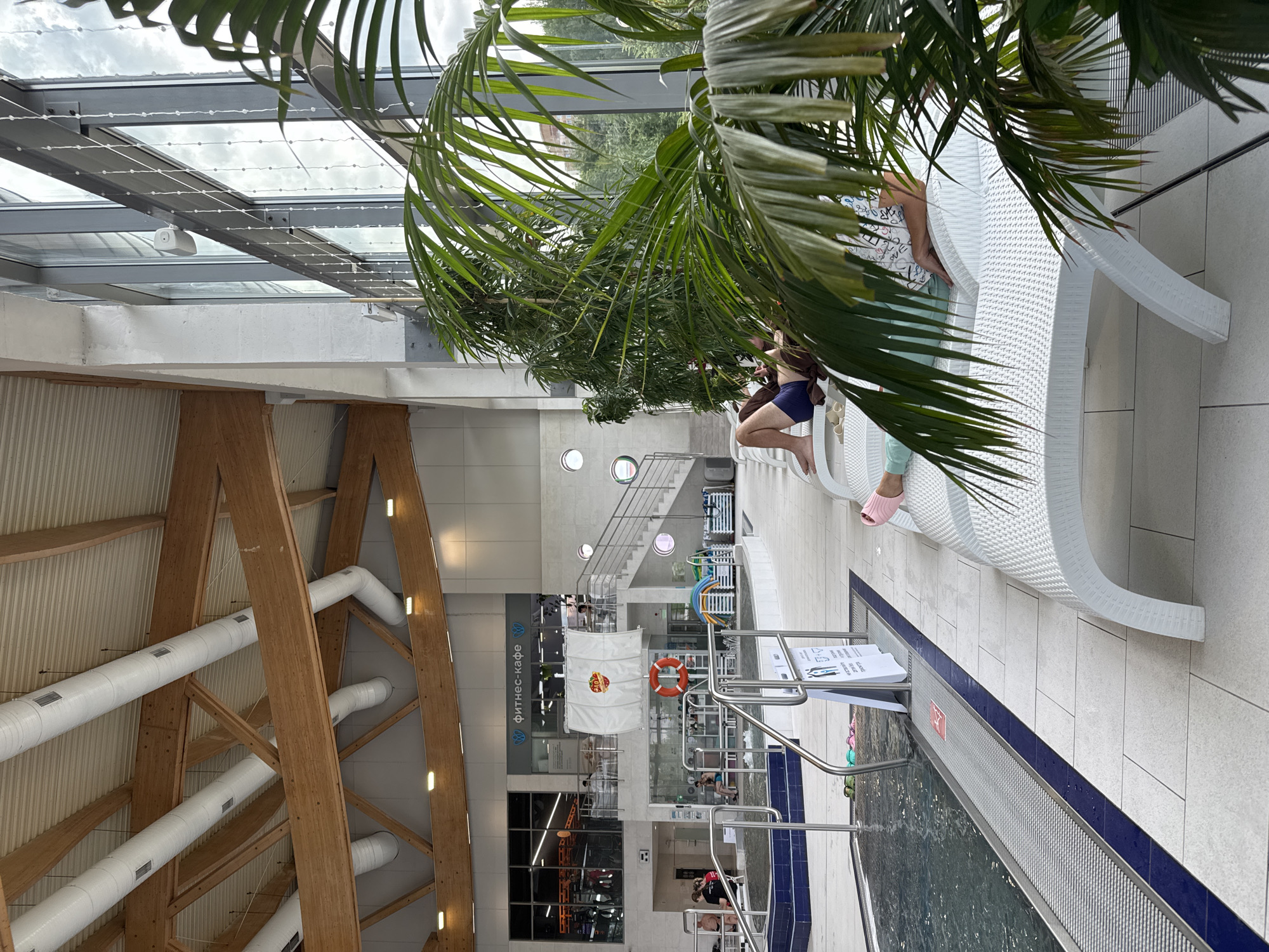 The image depicts an indoor swimming pool facility located in Sovkhoza imeni Lenina, Russia. In the foreground, a large potted palm tree partially obscures the view of white plastic lounge chairs. Several people are seated or lying on these chairs. One person in the mid-ground is lying on a lounge chair, wearing dark blue swimming trunks and a brown towel draped over their shoulders. Another individual, partially visible, is also on a lounge chair, wearing a light-colored top with a pattern and teal leggings. The lounge chairs are arranged on a tiled floor.
To the left, a section of the swimming pool is visible with dark blue tiling along the edge. There are metal handrails leading into the pool, and a sign with pictograms is posted nearby. Further back, a staircase with metal railings ascends, leading to a level where a sign for "фитнес-кафе" (fitness cafe) is displayed. Behind the fitness cafe sign, a white banner with a red and yellow circular logo hangs. In the background, the ceiling features exposed wooden beams and white ventilation ducts. Large windows line one side of the building, allowing natural light to enter. Other amenities like inflatable toys and chairs are visible on a platform beyond the pool.