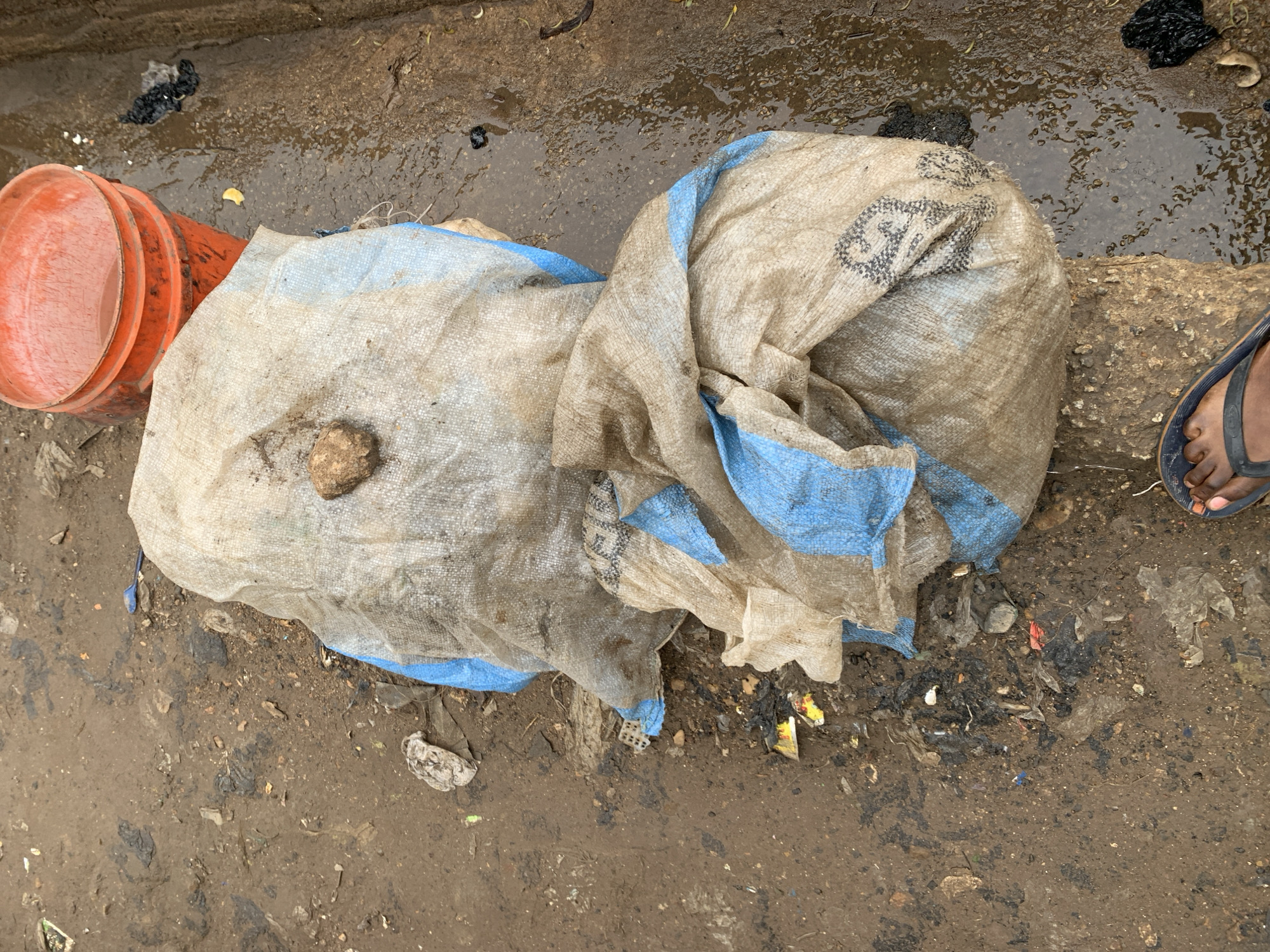 The image shows a ground-level view of what appears to be a street or roadside in Rafin Par, Nigeria. The ground is muddy and littered with debris. Two large woven sacks, dirty and worn, occupy the center of the frame. One orange plastic bucket is visible to the left of the sacks. A foot wearing a sandal is visible to the right. The background shows wet pavement or concrete.